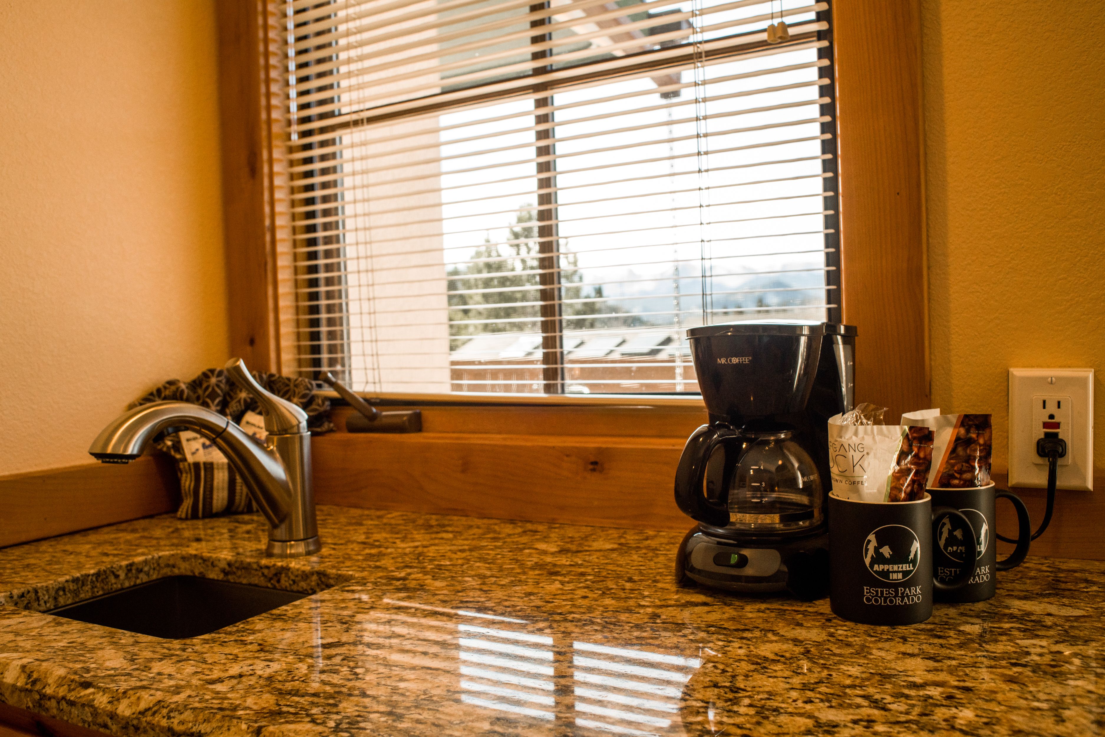 Una encimera de cocina con una cafetera, un lavabo y una ventana con persianas.