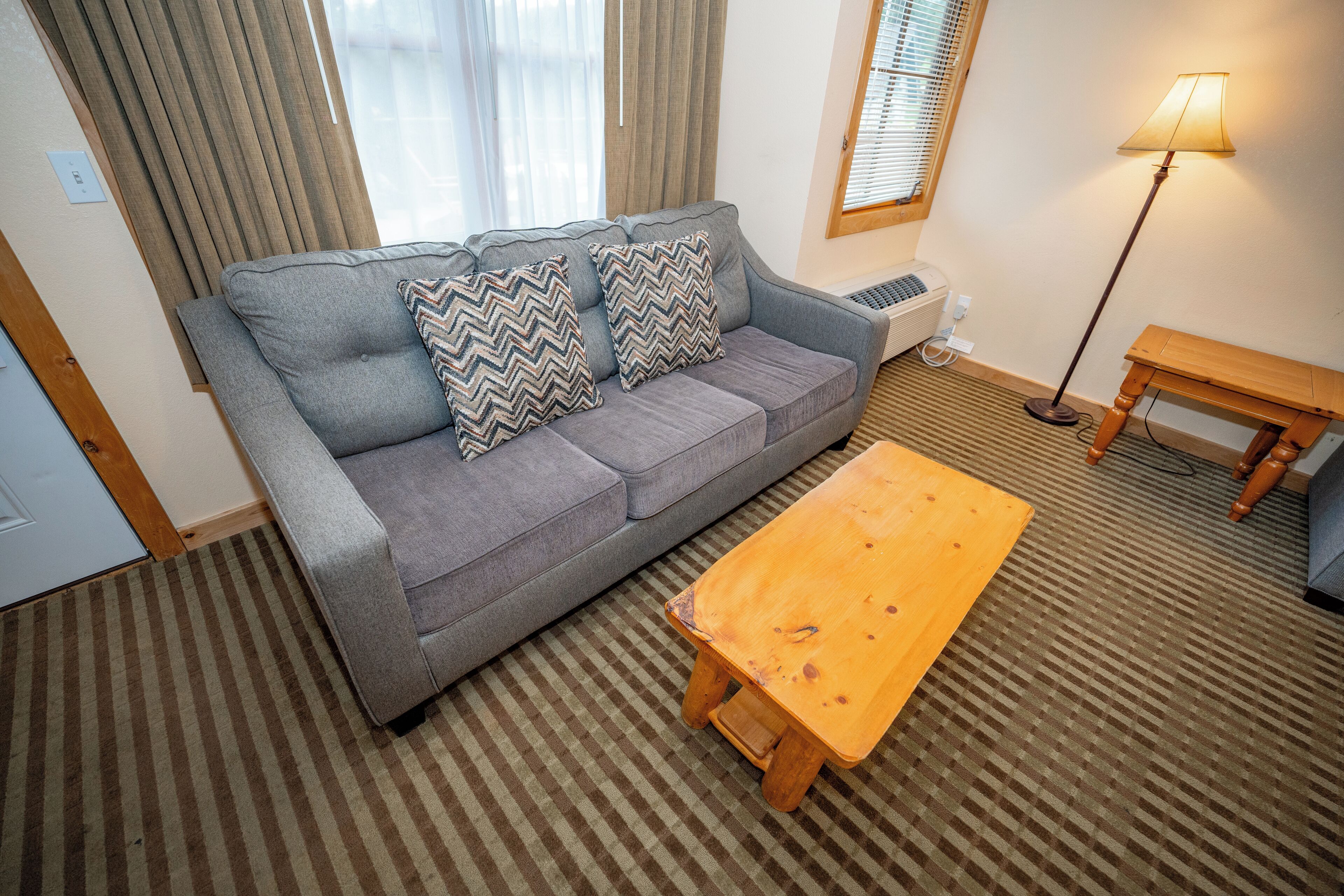 A living room with a grey sofa, patterned cushions, a wooden coffee table, a standing lamp, and a striped carpet.
