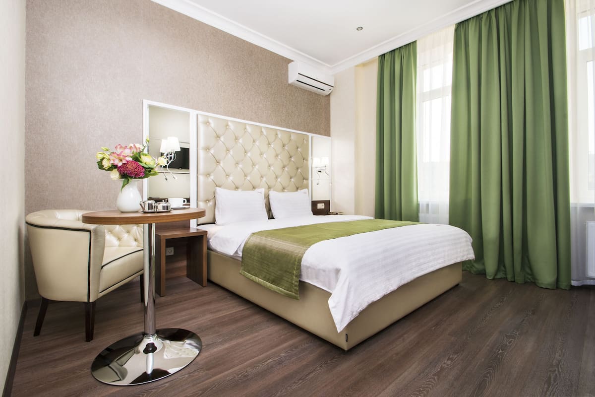 A hotel room with a bed, a vanity with a mirror, a chair, a table with a vase of flowers, and a window with green curtains.