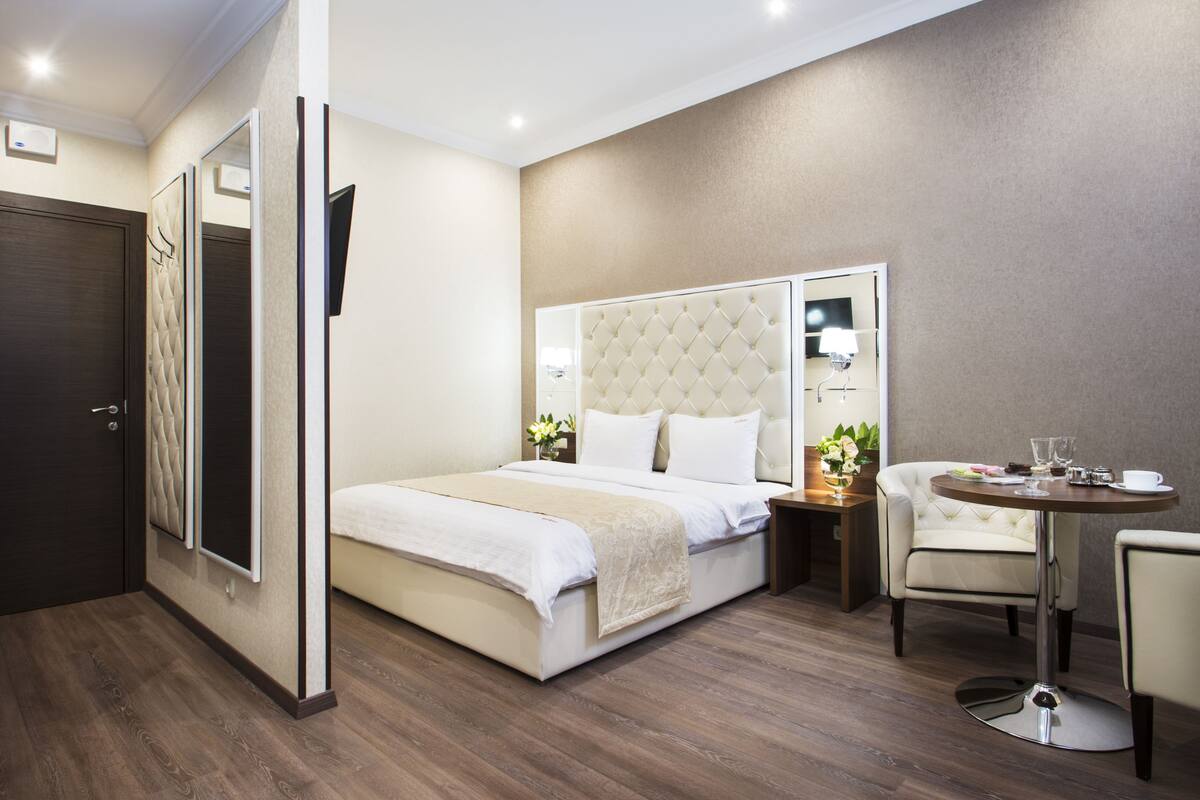 A modern hotel room with a large bed, a round table, a chair, and a small side table with a lamp.