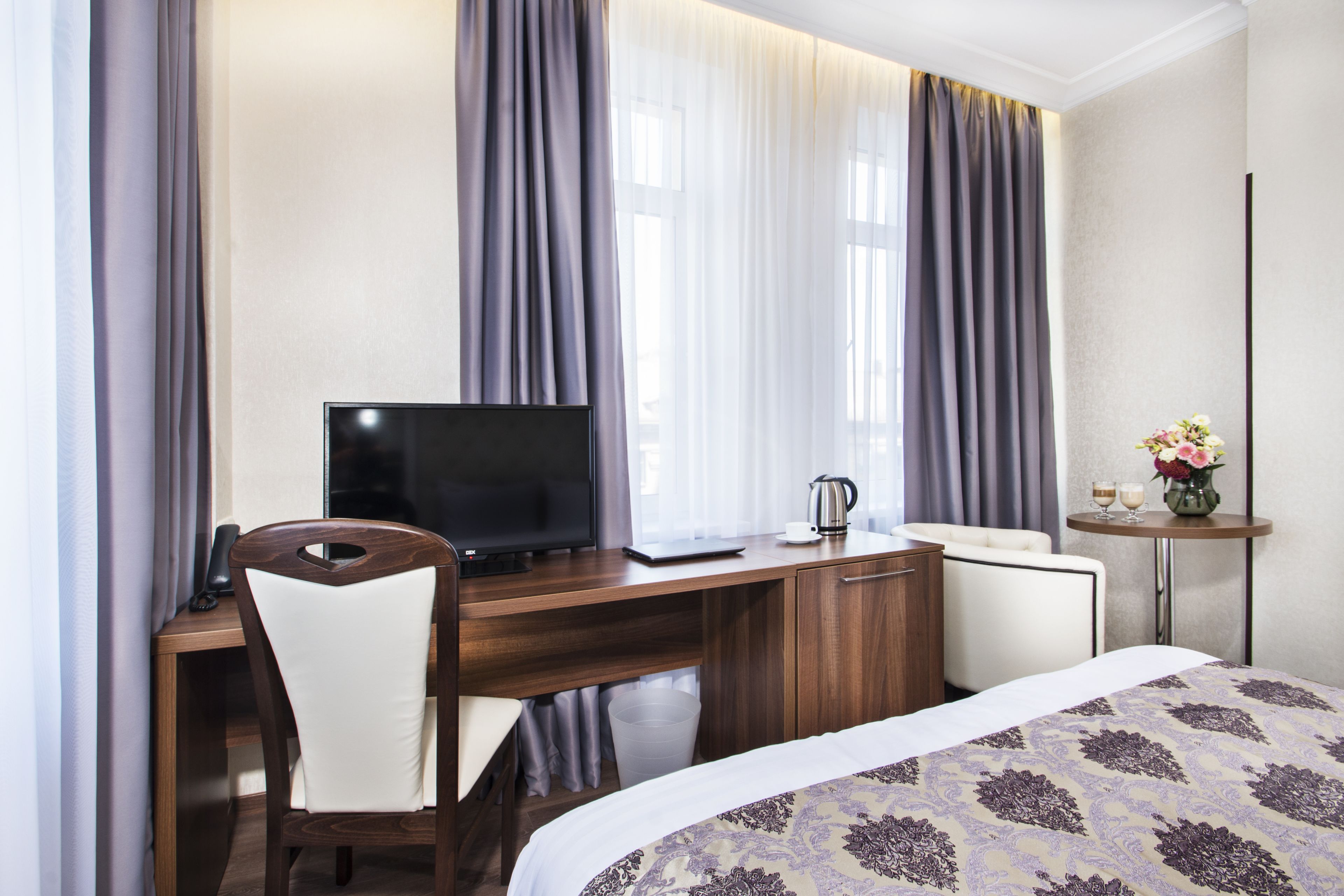 A hotel room with a wooden desk, a television, a laptop, a chair, a bed with a patterned bedspread, a small table with a vase of flowers, and curtains.