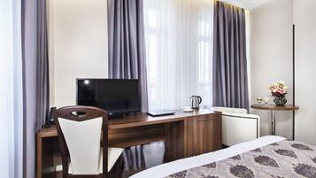 A hotel room with a wooden desk, a television, a laptop, a chair, a bed with a patterned bedspread, a small table with a vase of flowers, and curtains.