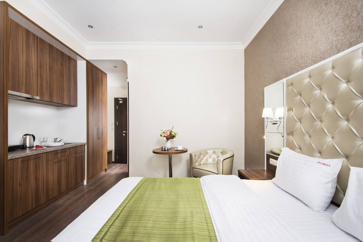 A hotel room with a bed, a small table with a vase of flowers, a chair, a minibar, and wooden cabinets.