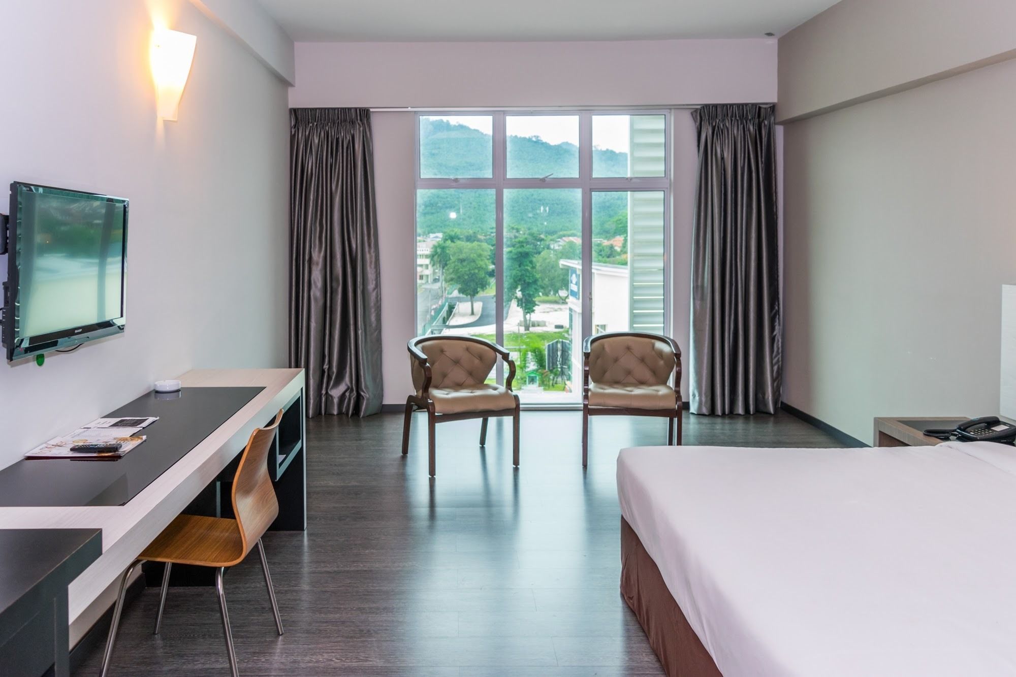 A hotel room with a bed, a desk with a chair, a TV, and a view of a cityscape through large windows.