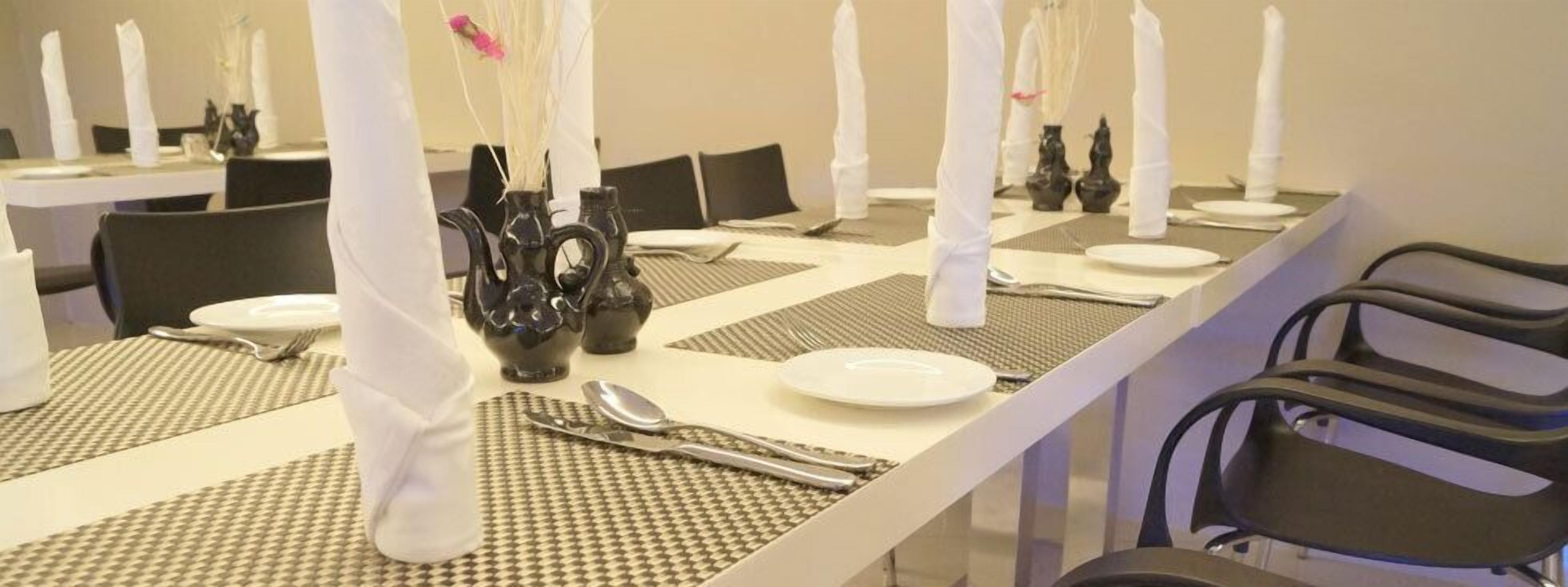 A dining area with tables set for meals, each with a place setting including a plate, cutlery, and a vase with flowers.