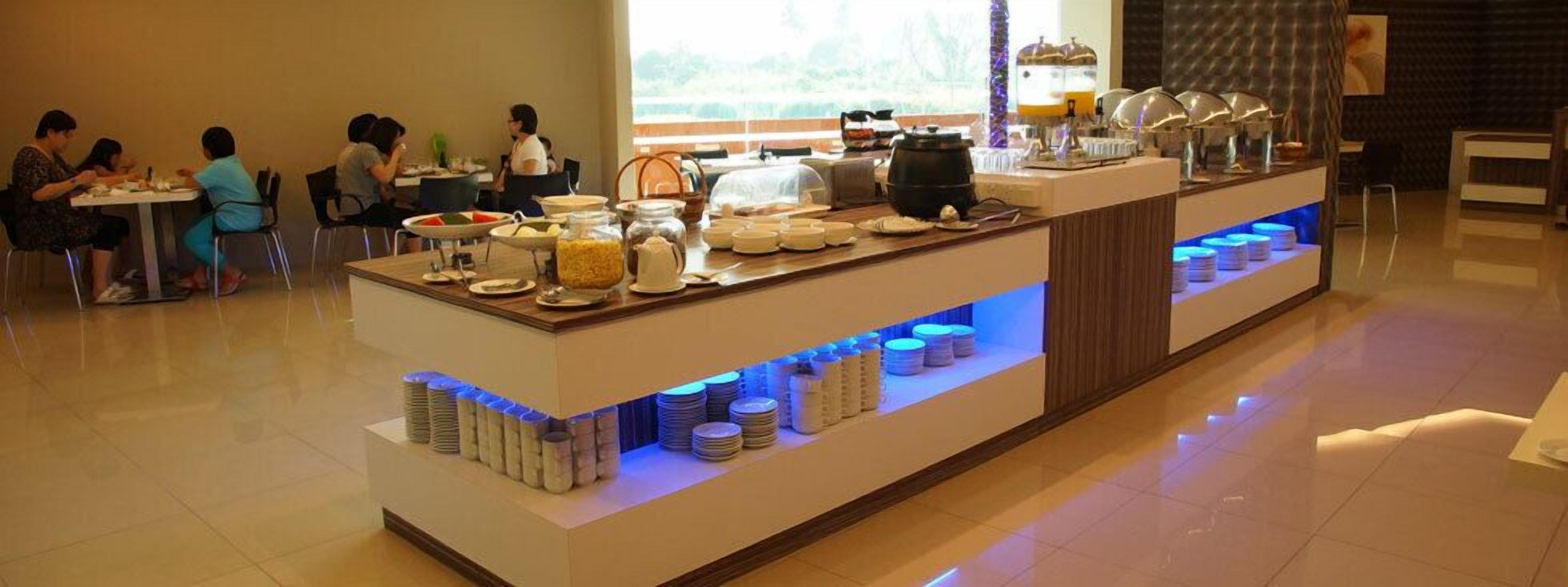 A buffet setup with various food items and utensils, a coffee machine, and a seating area with people dining.