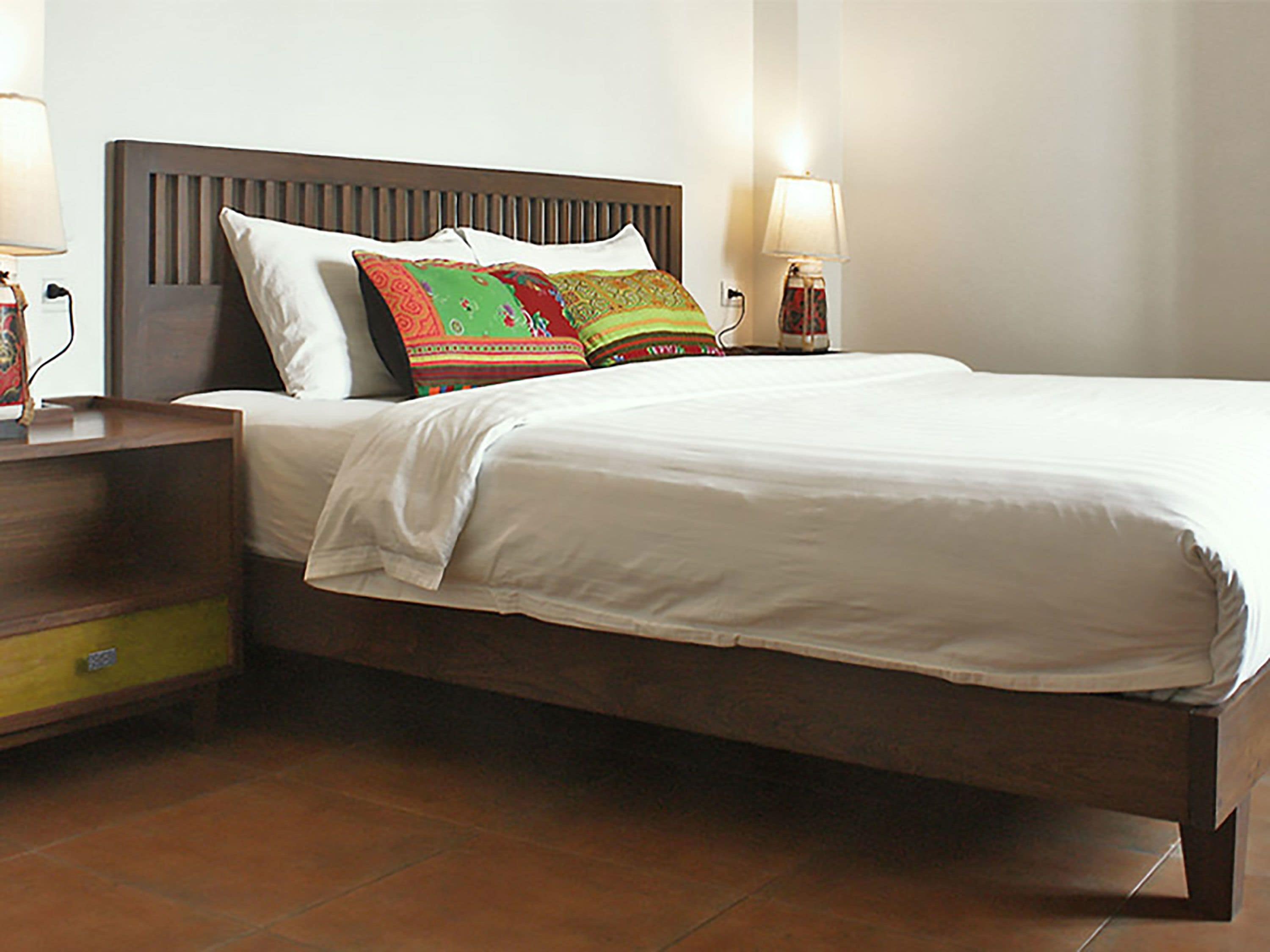 Cama bem arrumada com lençóis brancos, cabeceira de madeira e almofadas coloridas. Há uma mesa de cabeceira com uma luminária e um pequeno objeto decorativo. O quarto tem piso de azulejo e uma parede lisa.