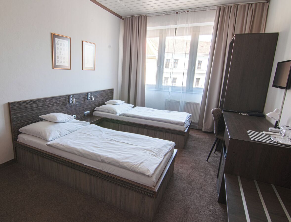 A hotel room with two single beds, a desk with a chair, a television, and a window with curtains.