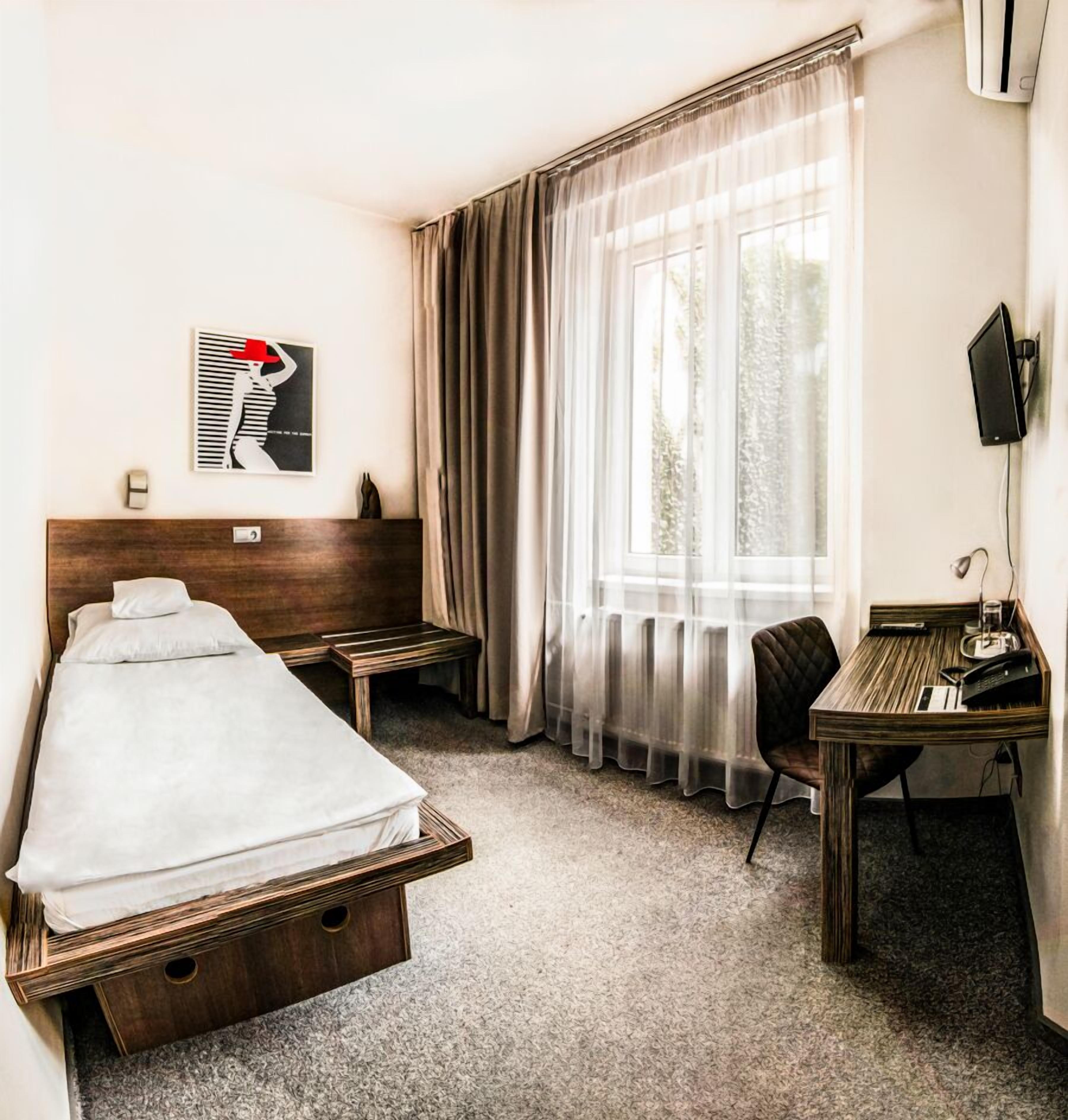 A hotel room with a single bed, a desk with a chair, a television, and a window with sheer curtains.