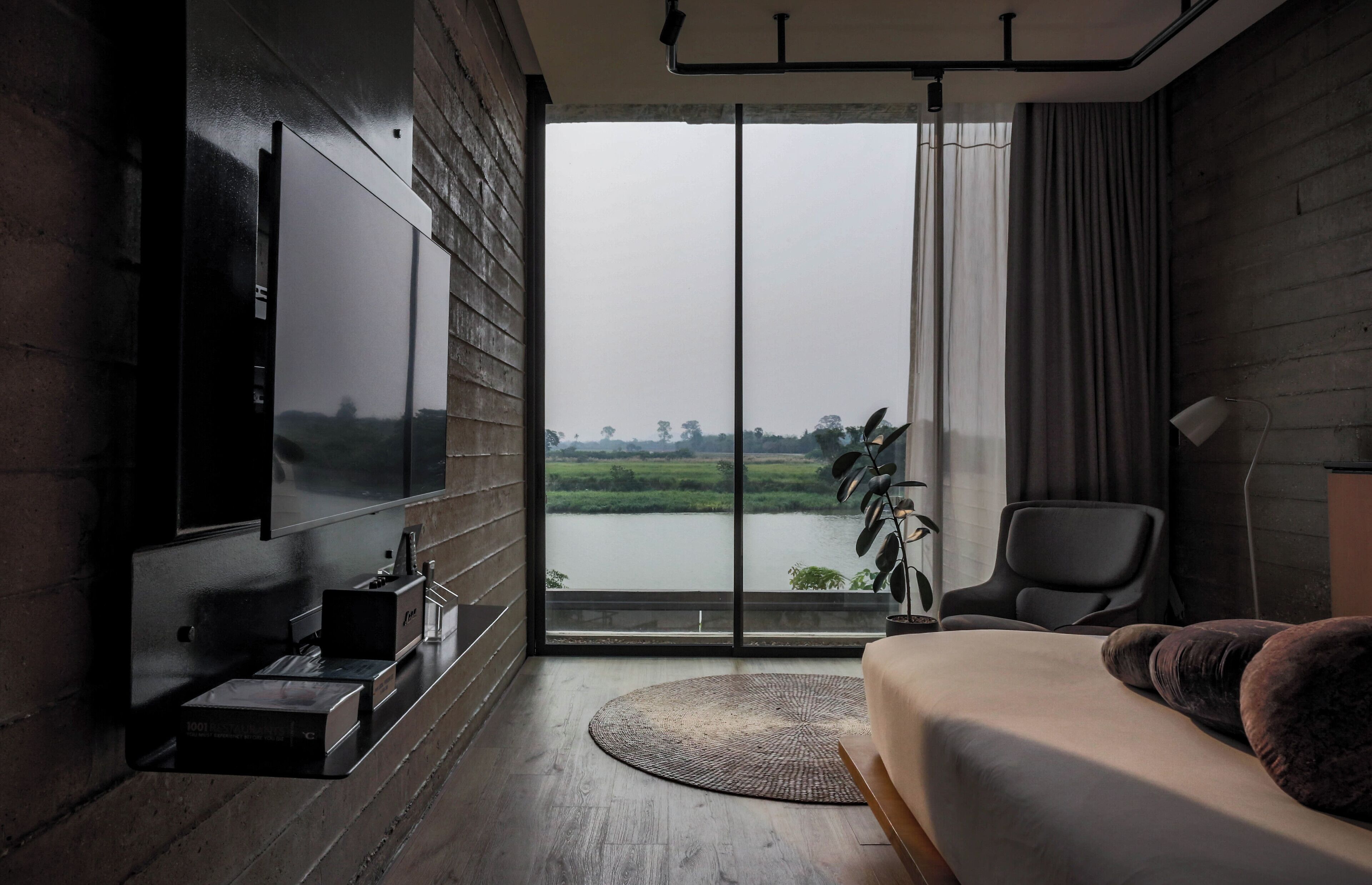 A modern bedroom with a large TV, a bed, a chair, and a view of a lake through a large window.