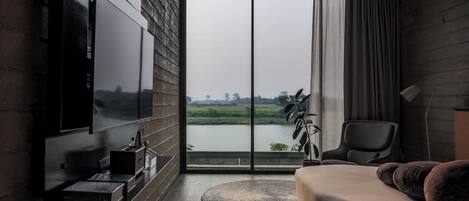 A modern bedroom with a large TV, a bed, a chair, and a view of a lake through a large window.