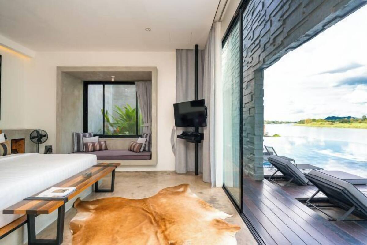 Ein modernes Schlafzimmer mit Bett, einer Holzbank, einem Fernseher und einem großen Fenster mit Blick auf einen Pool.