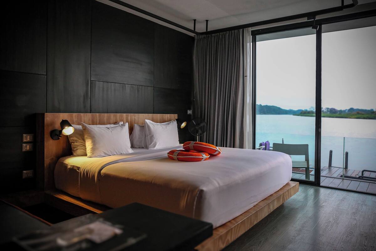 Ein modernes Hotelzimmer mit einem großen Bett, einem hölzernen Kopfteil, einem Nachttisch und Blick auf ein Gewässer durch große Fenster.
