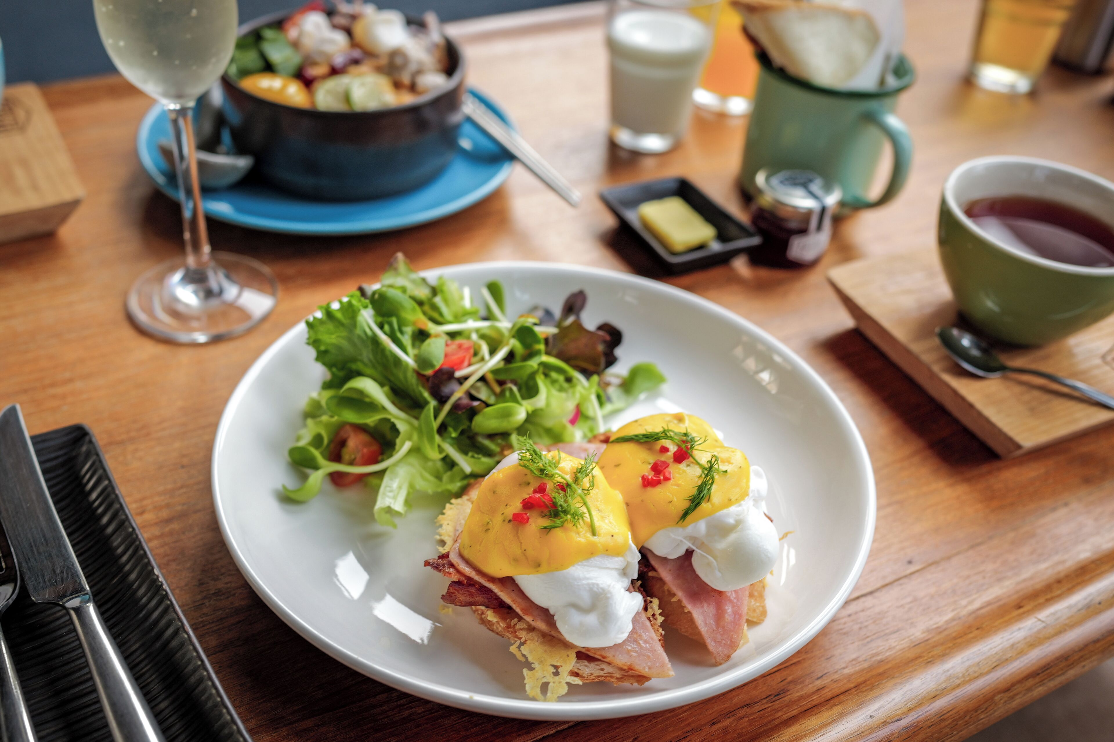Ein Frühstücksteller mit Eggs Benedict, einer grünen Tasse Tee und einer Salatschüssel auf einem Holztisch.