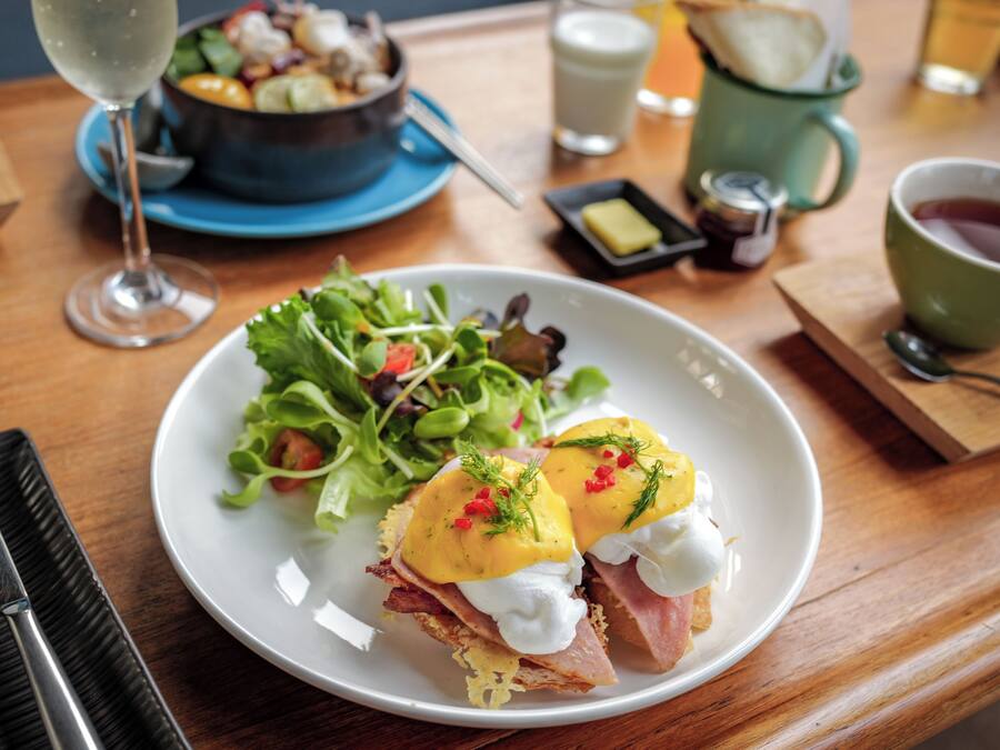 Ein Frühstücksteller mit Eggs Benedict, einer grünen Tasse Tee und einer Salatschüssel auf einem Holztisch.