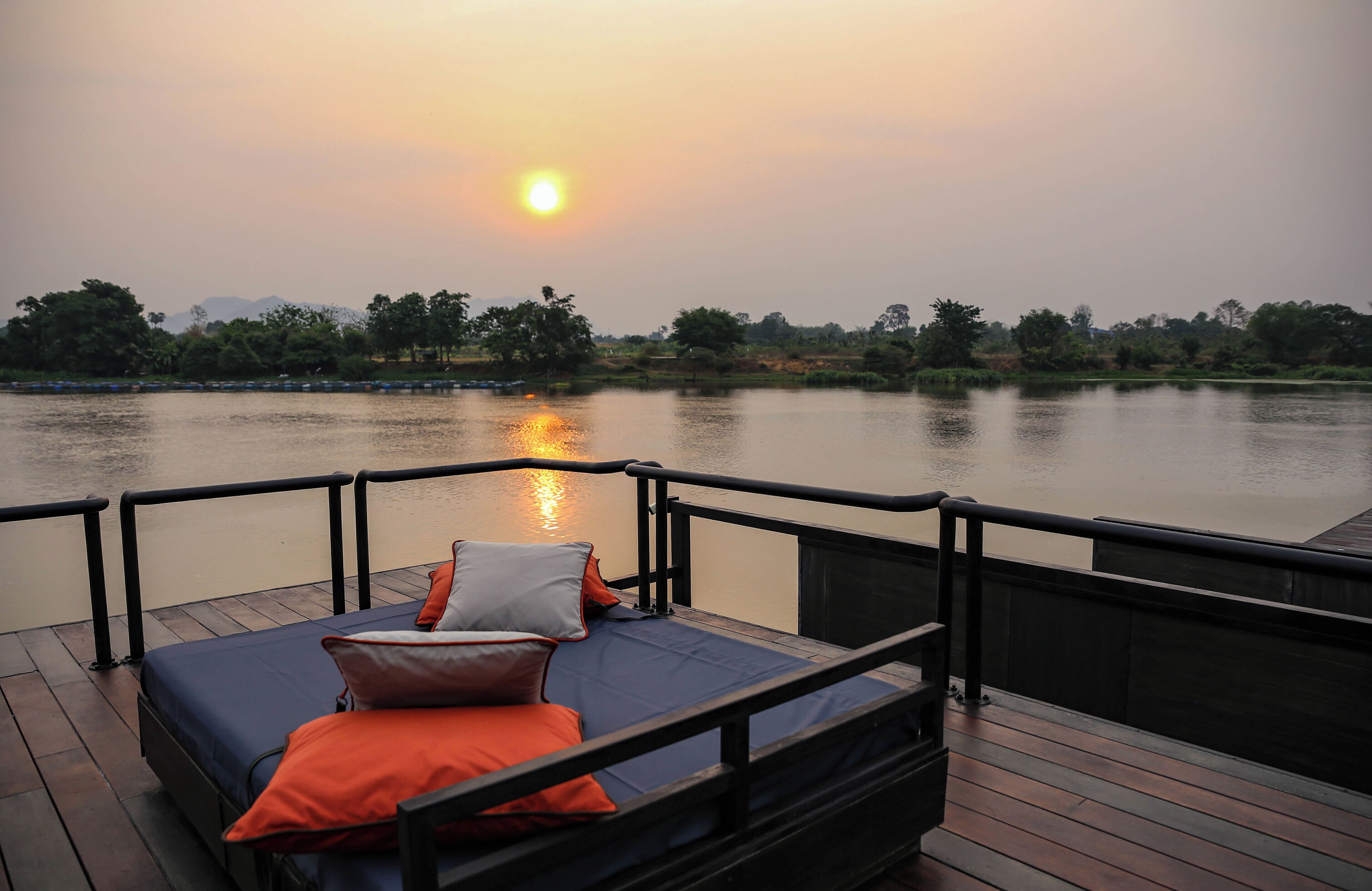 Een bed met kussens op een houten dek met uitzicht op een rustig wateroppervlak tijdens de zonsondergang.