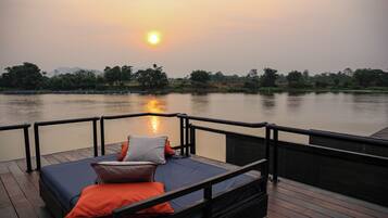 Een bed met kussens op een houten dek met uitzicht op een rustig wateroppervlak tijdens de zonsondergang.