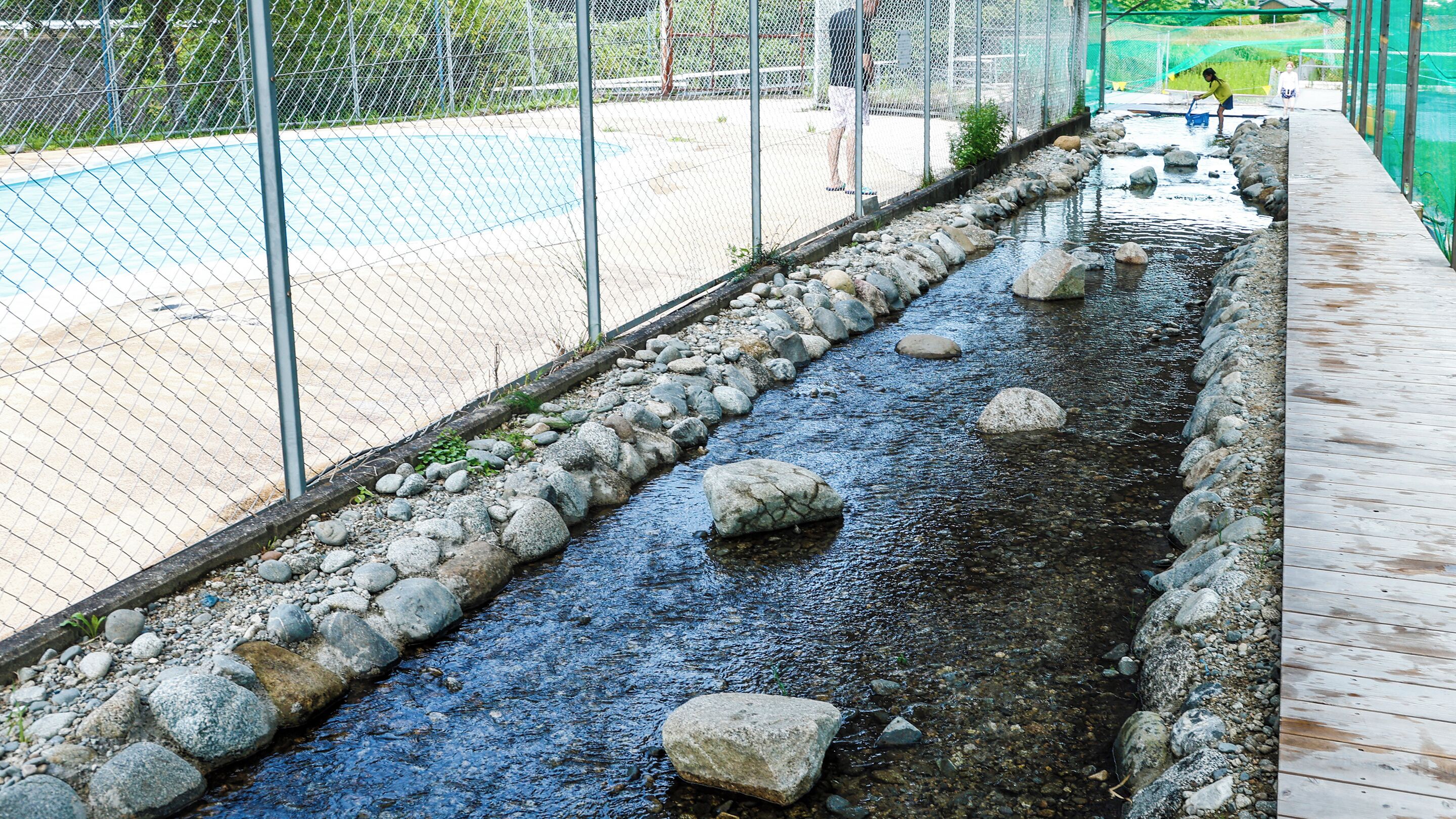 A water channel with rocks and a wooden walkway alongside a fenced area.