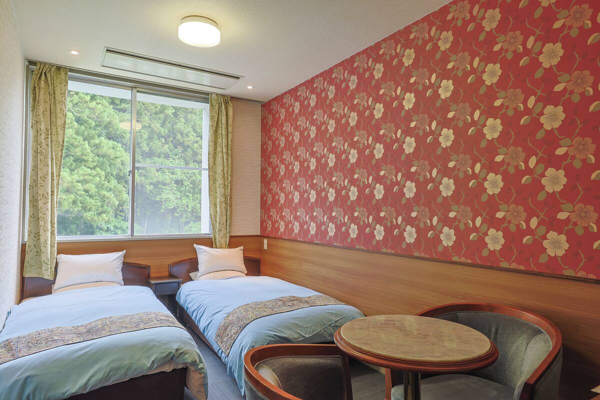 A hotel room with two beds, a small table, and a view of greenery through the window.