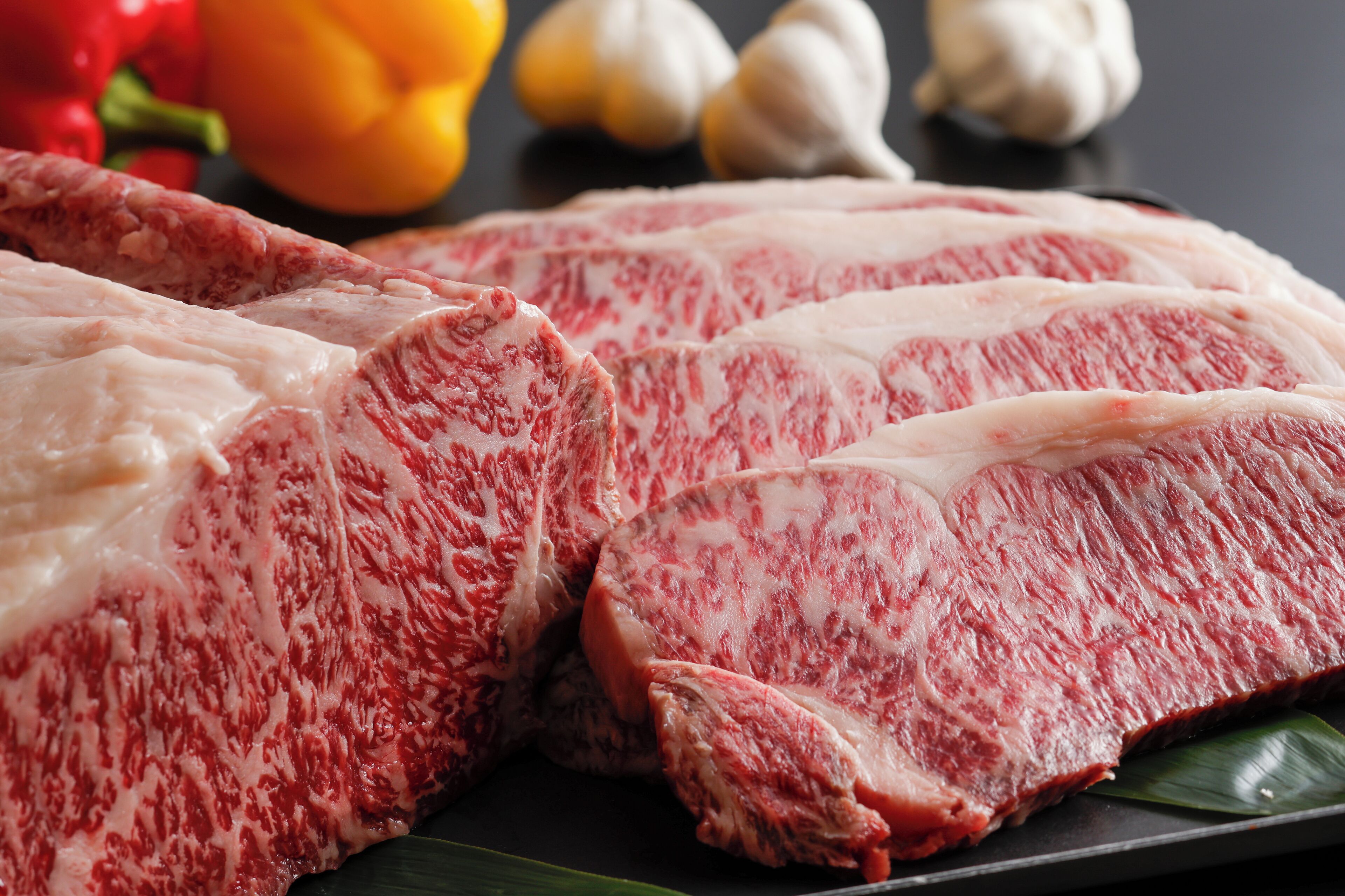 Raw meat cuts on a black surface with vegetables in the background.