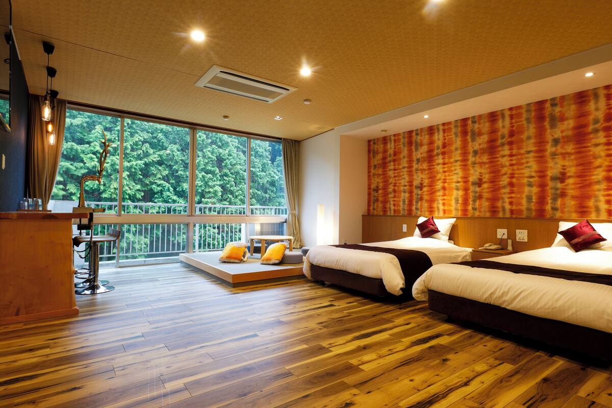 A hotel room with a large window, a wooden floor, two beds, a desk, and a balcony.