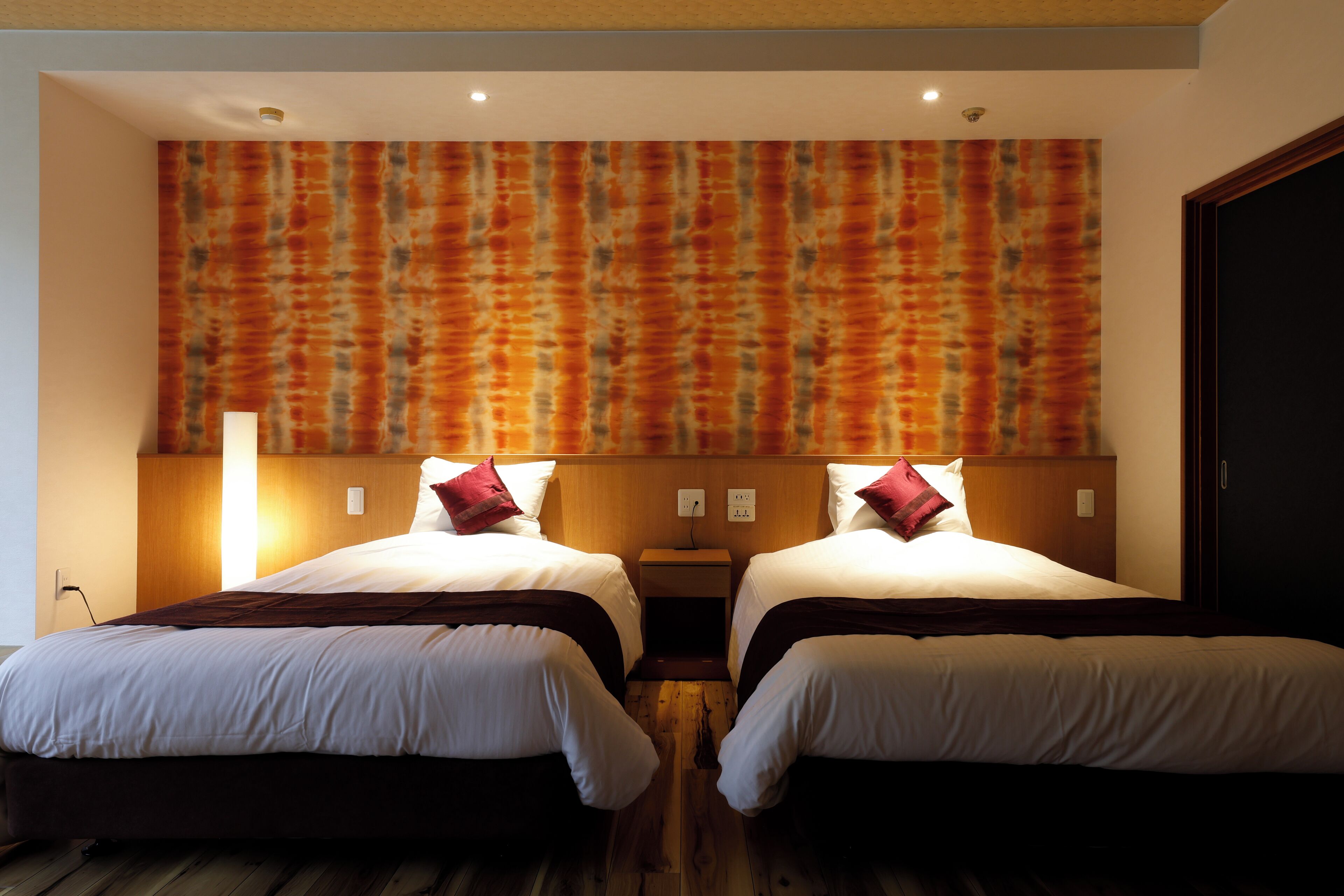 A hotel room with two beds, wooden flooring, a patterned wall, and a nightstand with a lamp.