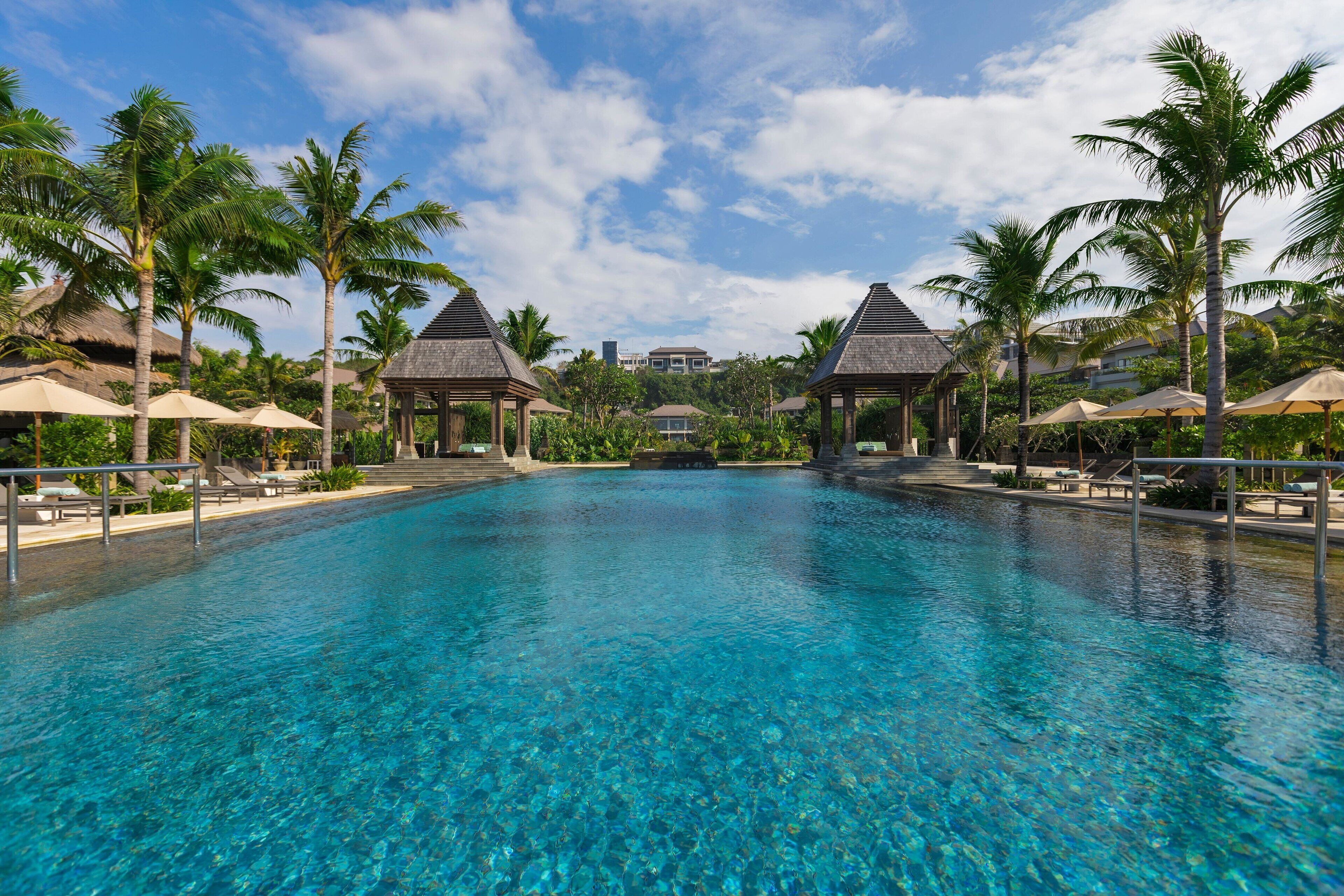 A clear blue pool with lounge chairs and umbrellas, surrounded by palm trees and a gazebo structure.