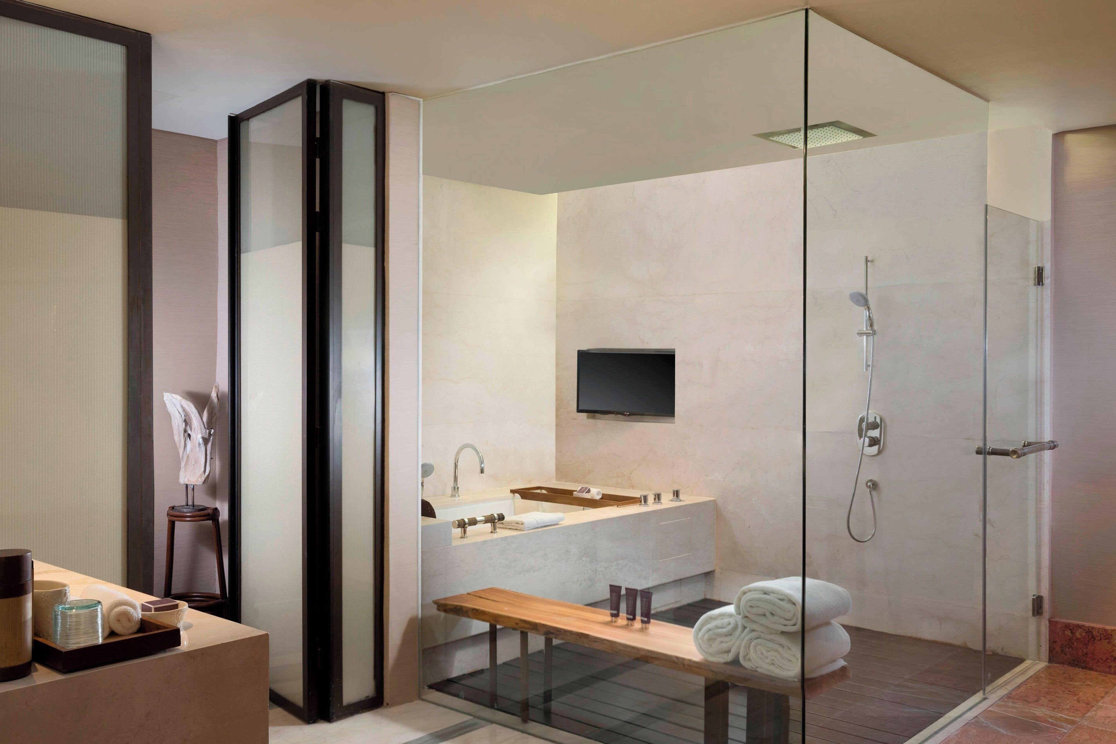 A modern bathroom with a glass shower enclosure, a wooden vanity, and a flat-screen TV mounted above the sink.