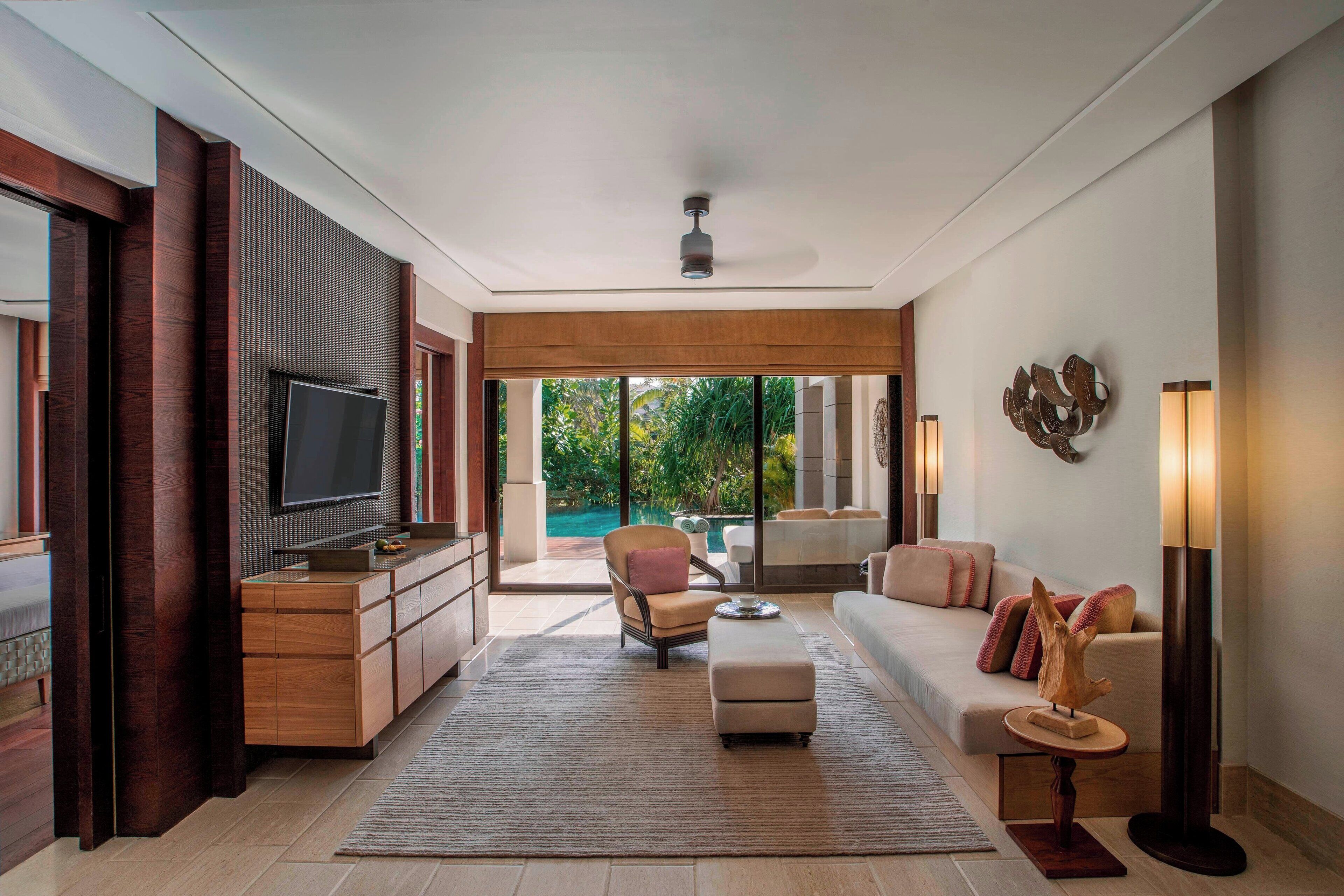 A modern living room with a TV, sofa, and armchair, a wooden cabinet, and a view of a pool and greenery.