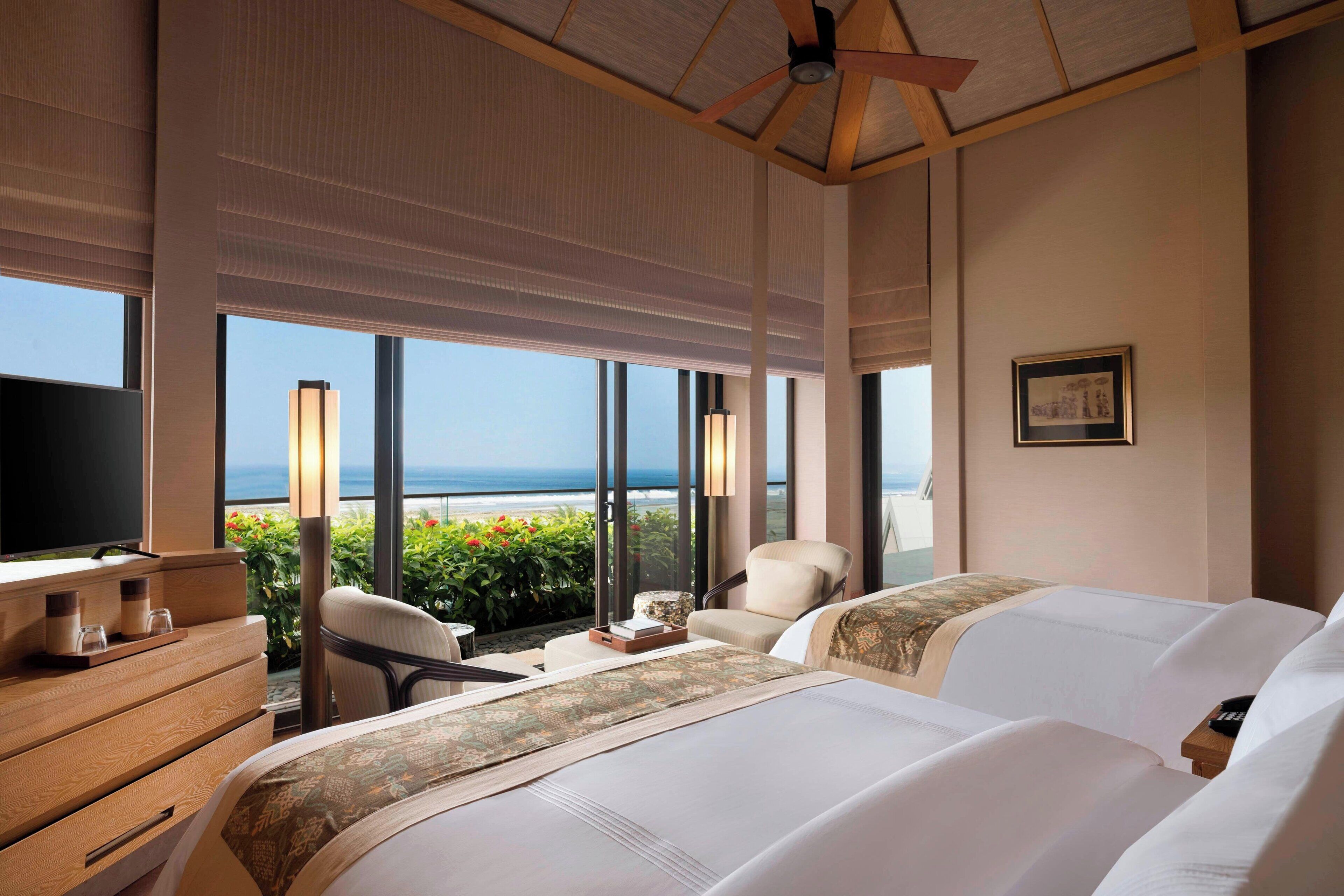 Habitación de hotel con dos camas, televisión, escritorio con lámpara y vista al océano.