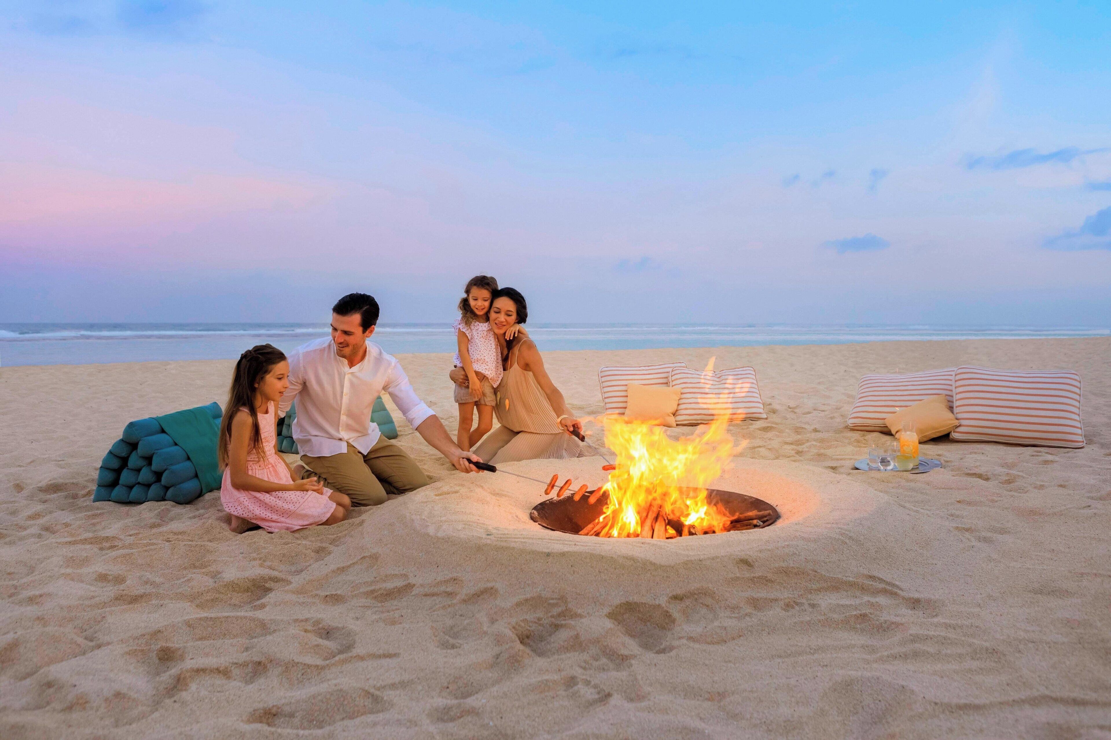 A family gathered around a campfire on a beach with a view of the ocean.