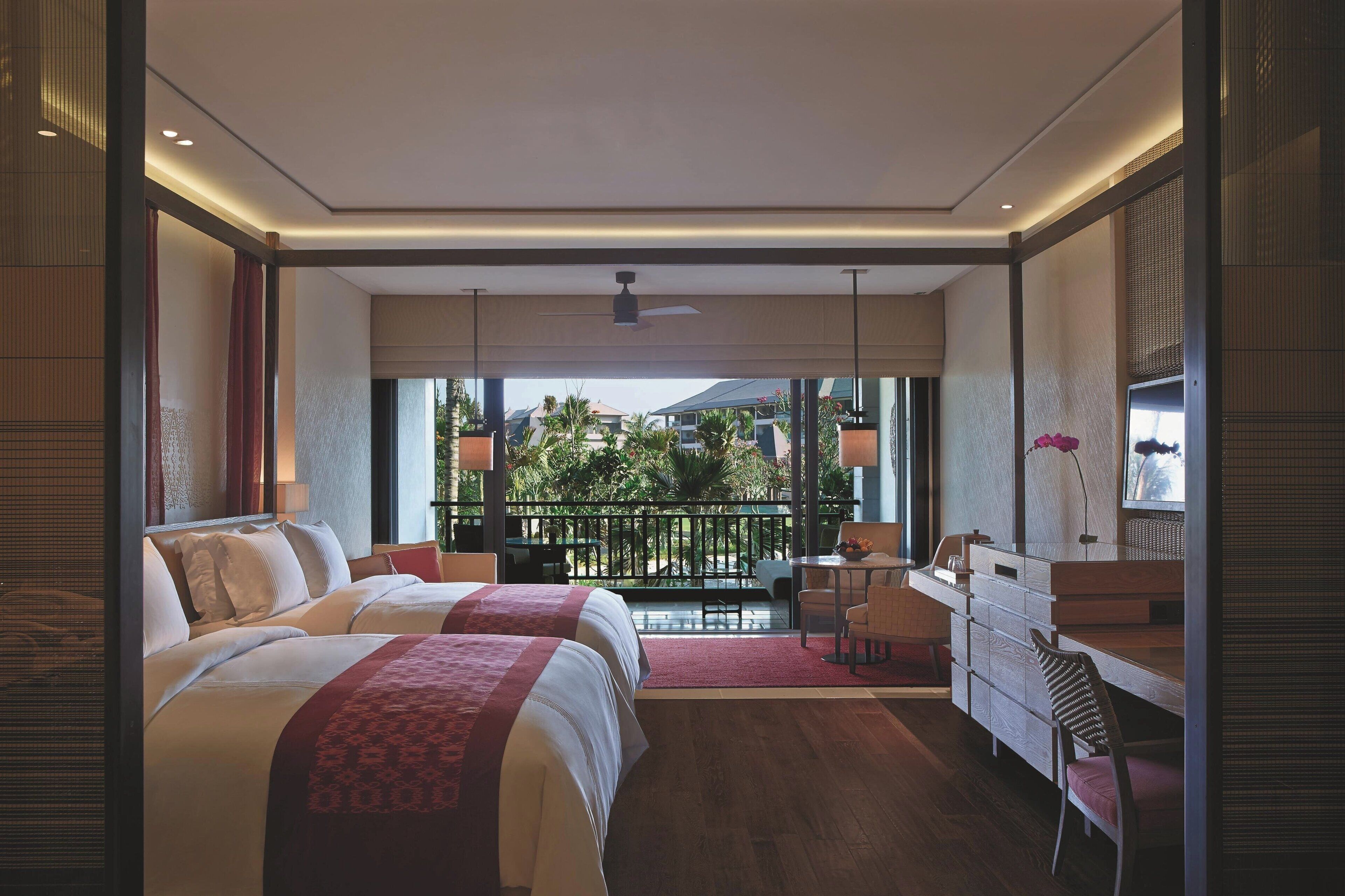 A hotel room with two beds, a desk, a chair, and a balcony with a view of greenery.
