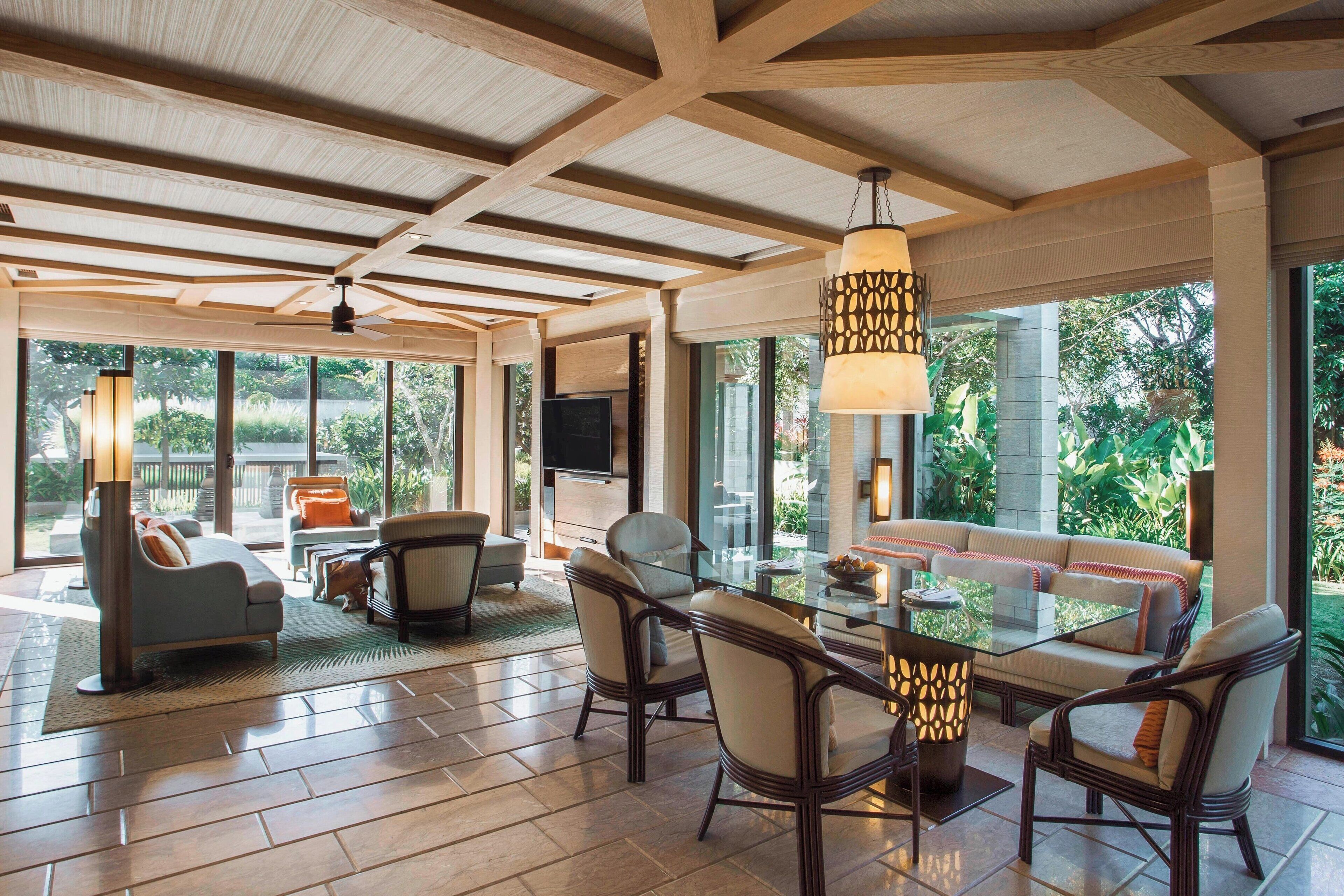 A spacious living area with a glass dining table, comfortable seating, and large windows offering a view of greenery.