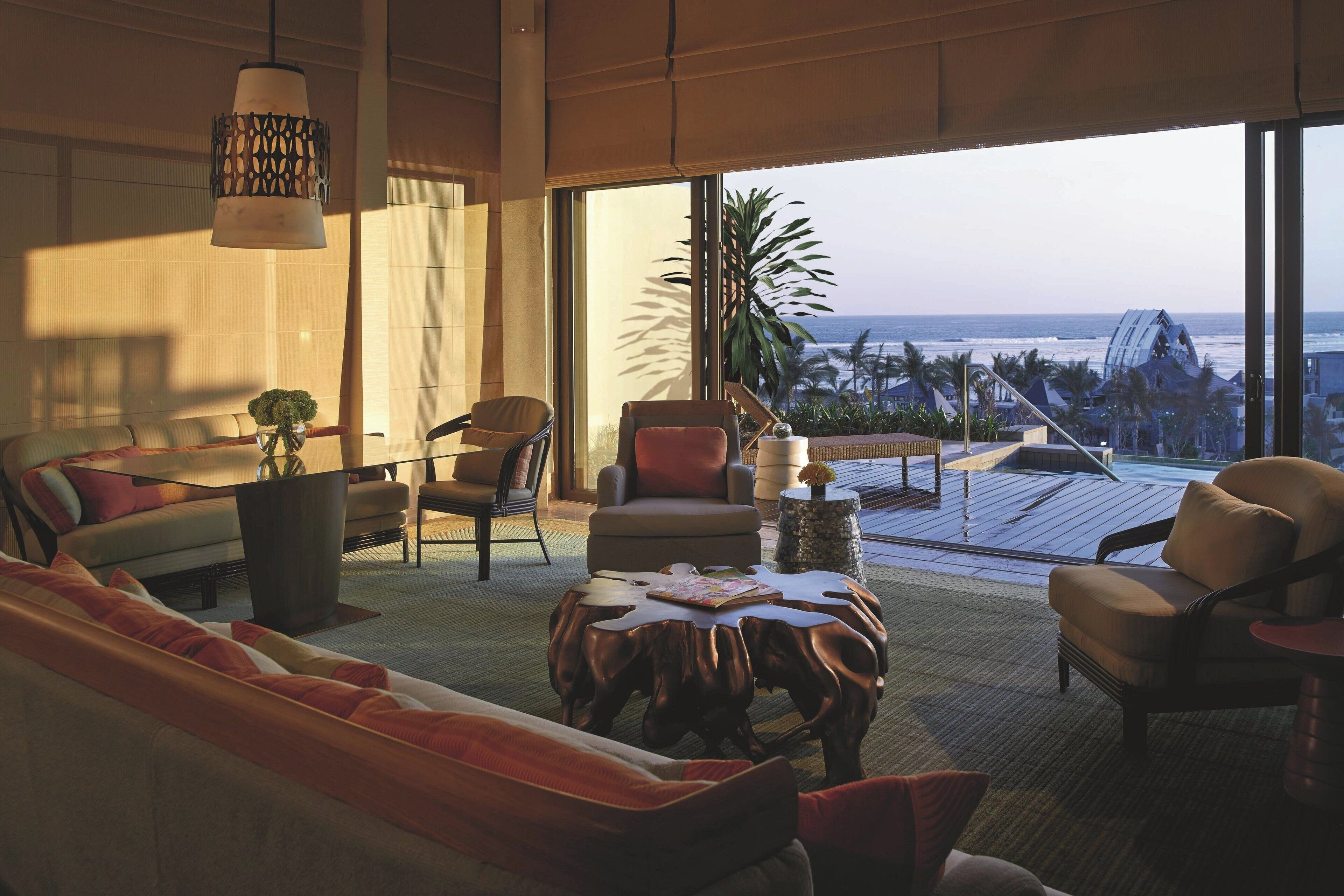 Sala de estar moderna com uma grande janela com vista para a praia, mobiliada com sofá, cadeiras e mesa de centro.