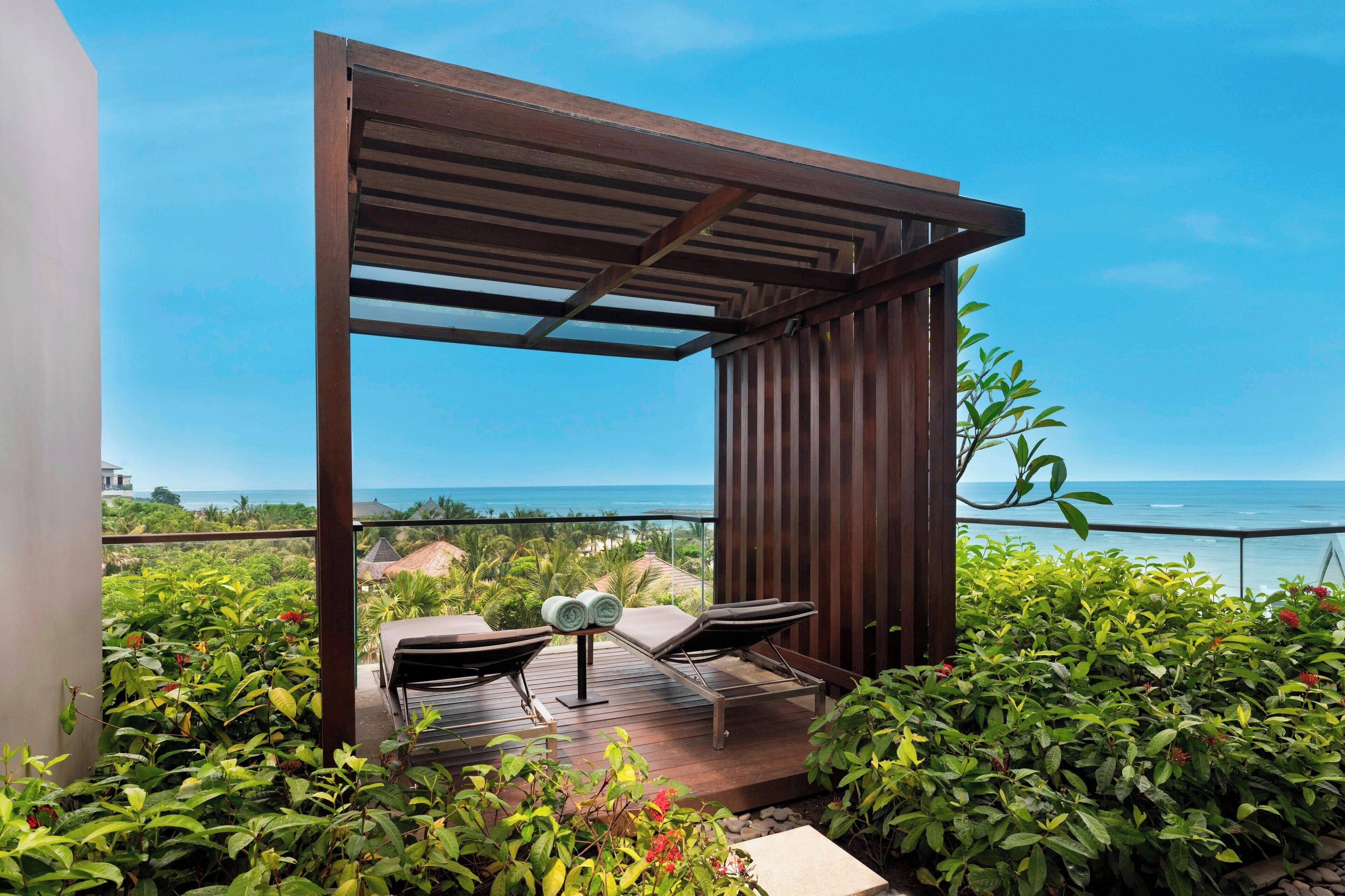 A wooden deck with two lounge chairs and a small table, overlooking a lush garden and the ocean.