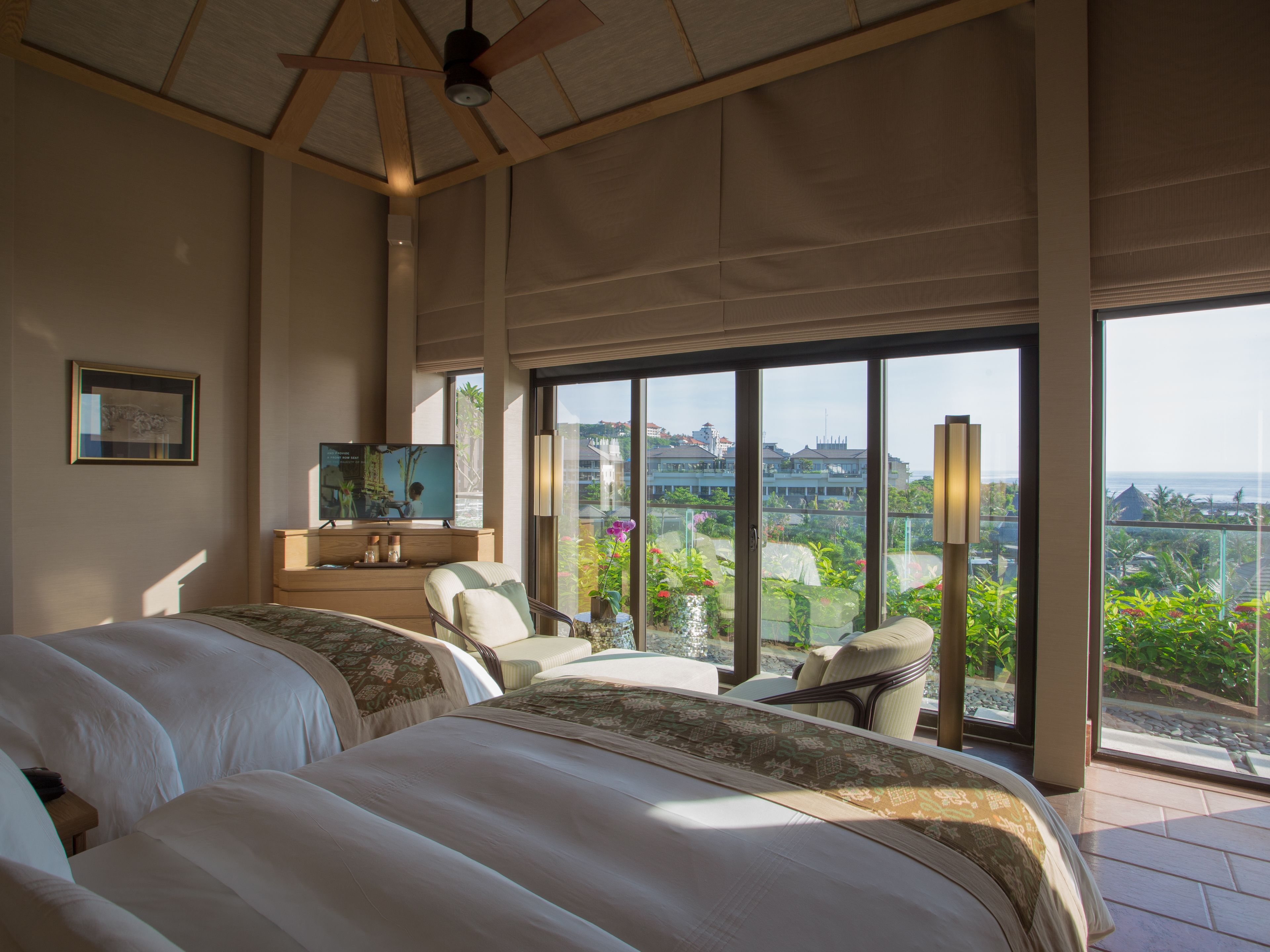 A spacious bedroom with two large beds, a television, and a balcony with a view of the ocean and buildings.