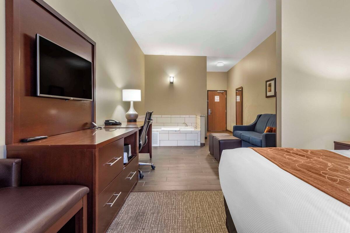 A hotel room with a bed, a desk with a TV, a chair, a sofa, and a bathroom with a bathtub.
