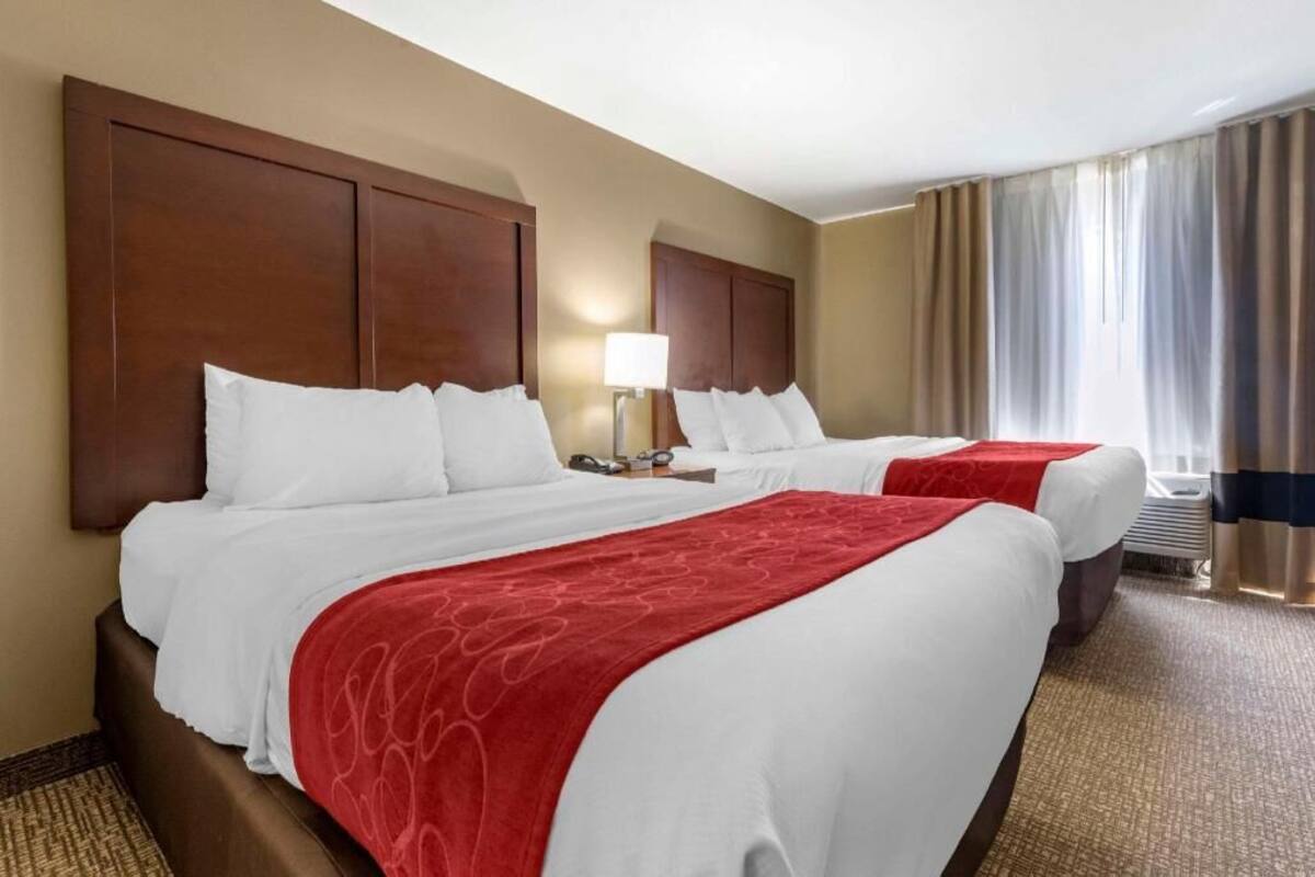 A hotel room with two beds, a large wooden headboard, a red throw blanket, a lamp, and a view of the outdoors.