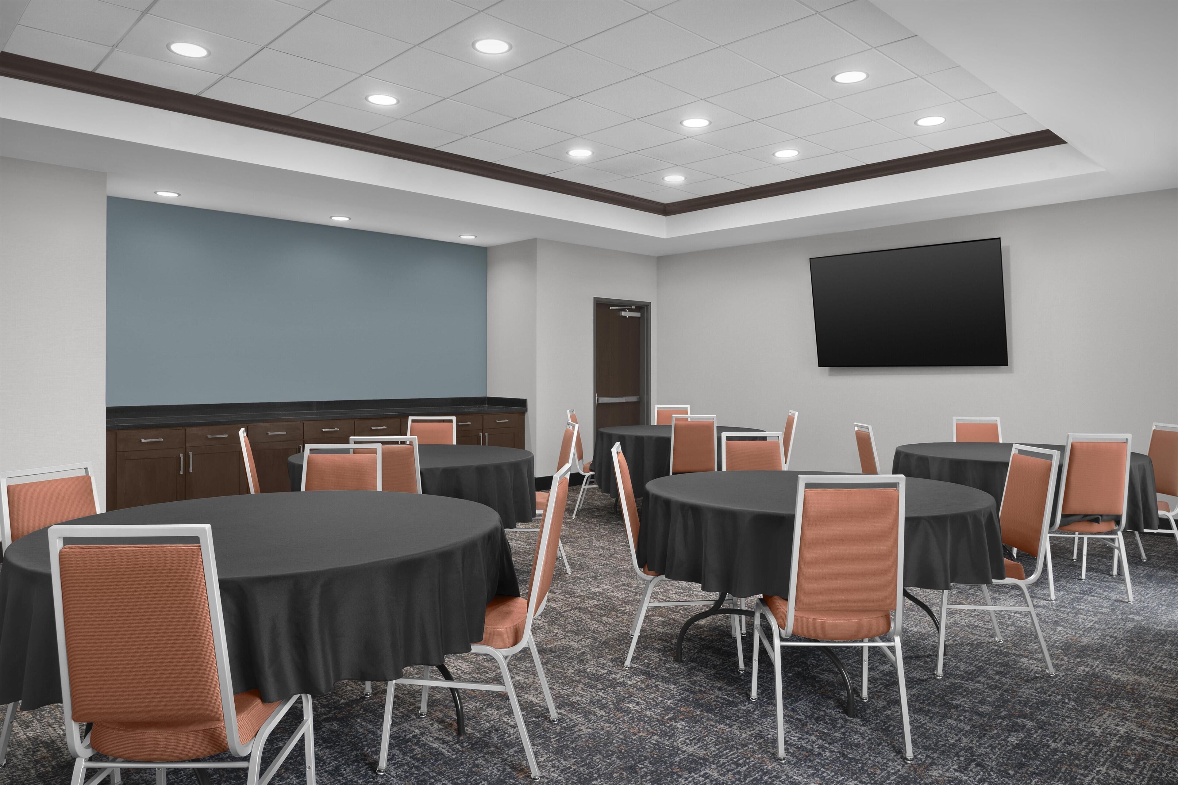 A conference room with multiple tables and chairs, a flat-screen TV on the wall, and a door in the background.