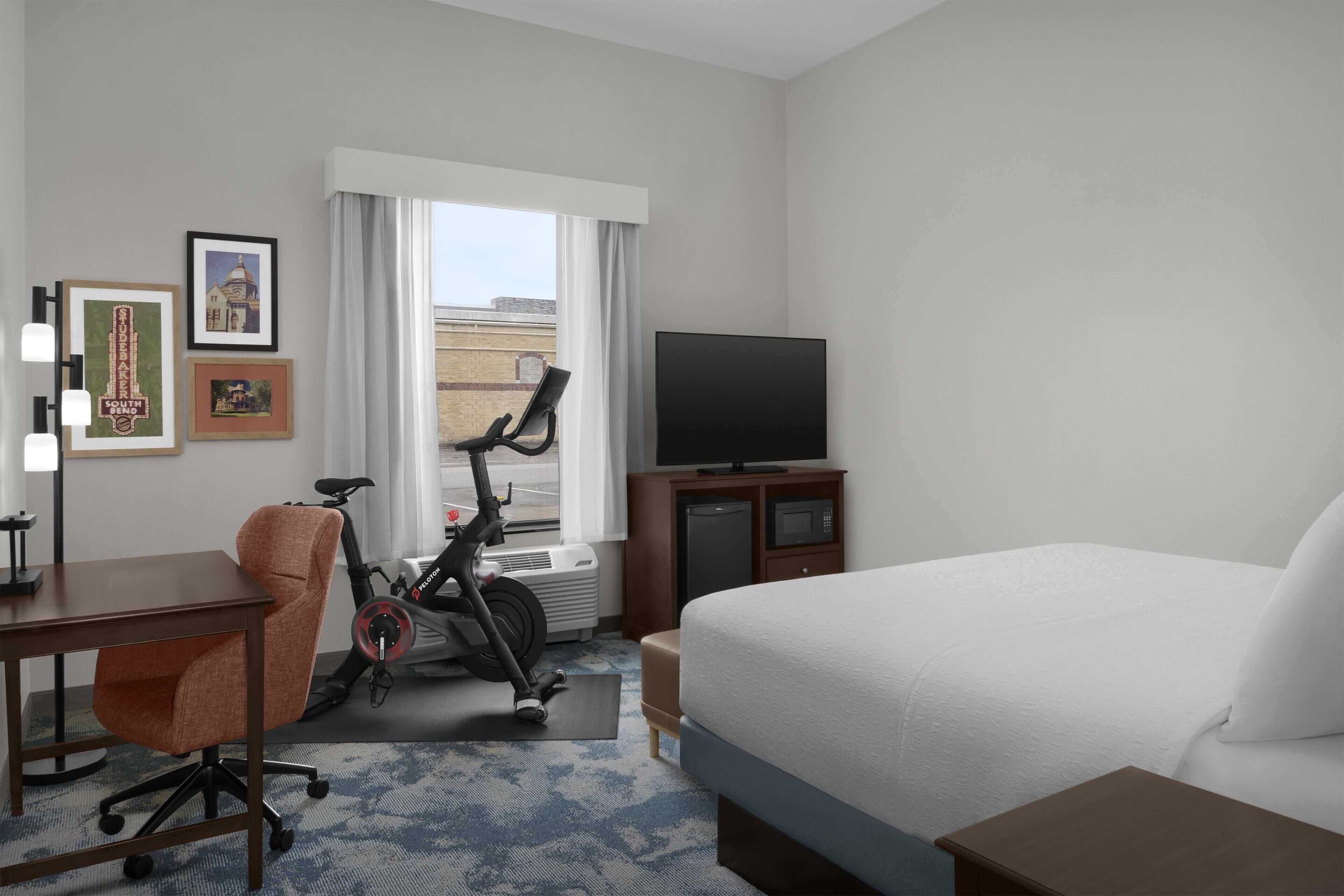 A hotel room with a bed, a desk, an exercise bike, a television, and framed pictures on the wall.