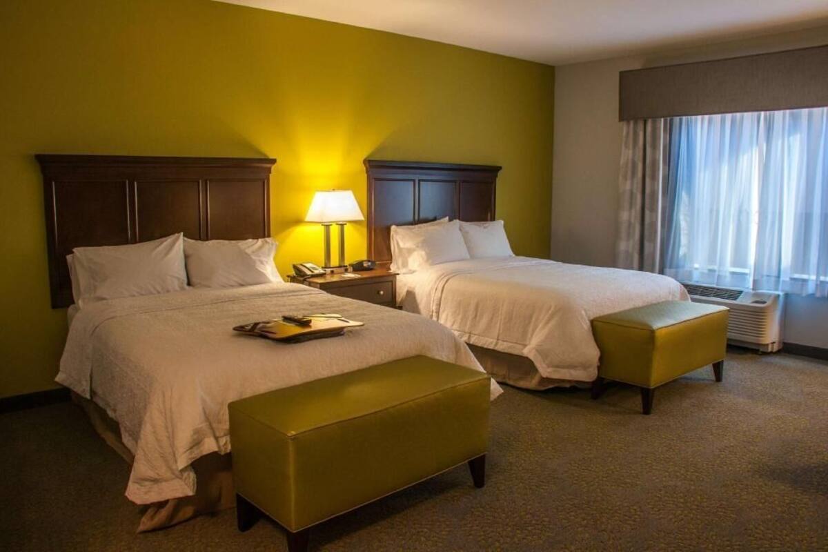 A hotel room with two beds, a bedside table with a lamp, an air conditioning unit, and a window with curtains.