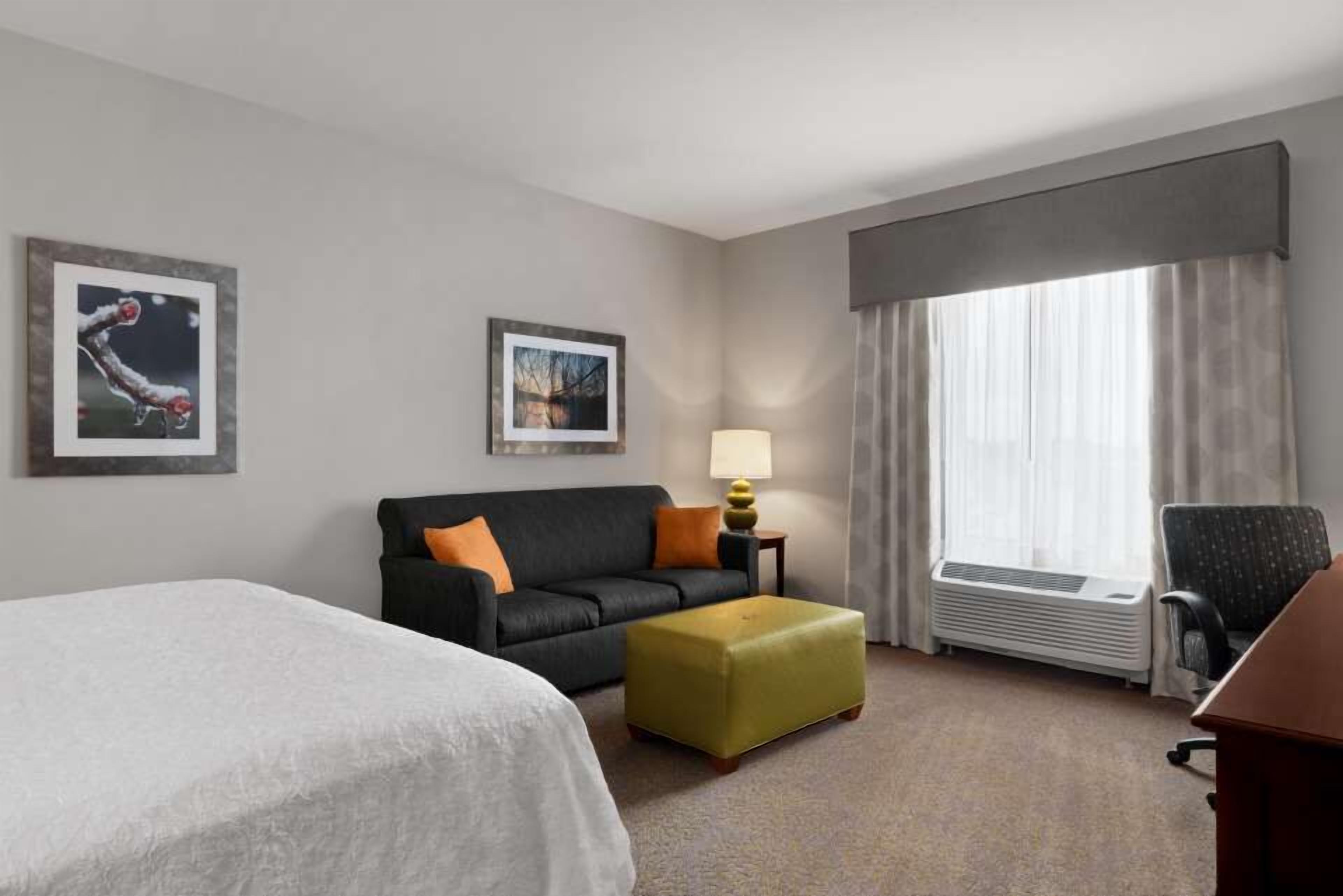 Habitación de hotel con cama, sofá, otoman, escritorio, lámpara, calefactor y dos cuadros en la pared.