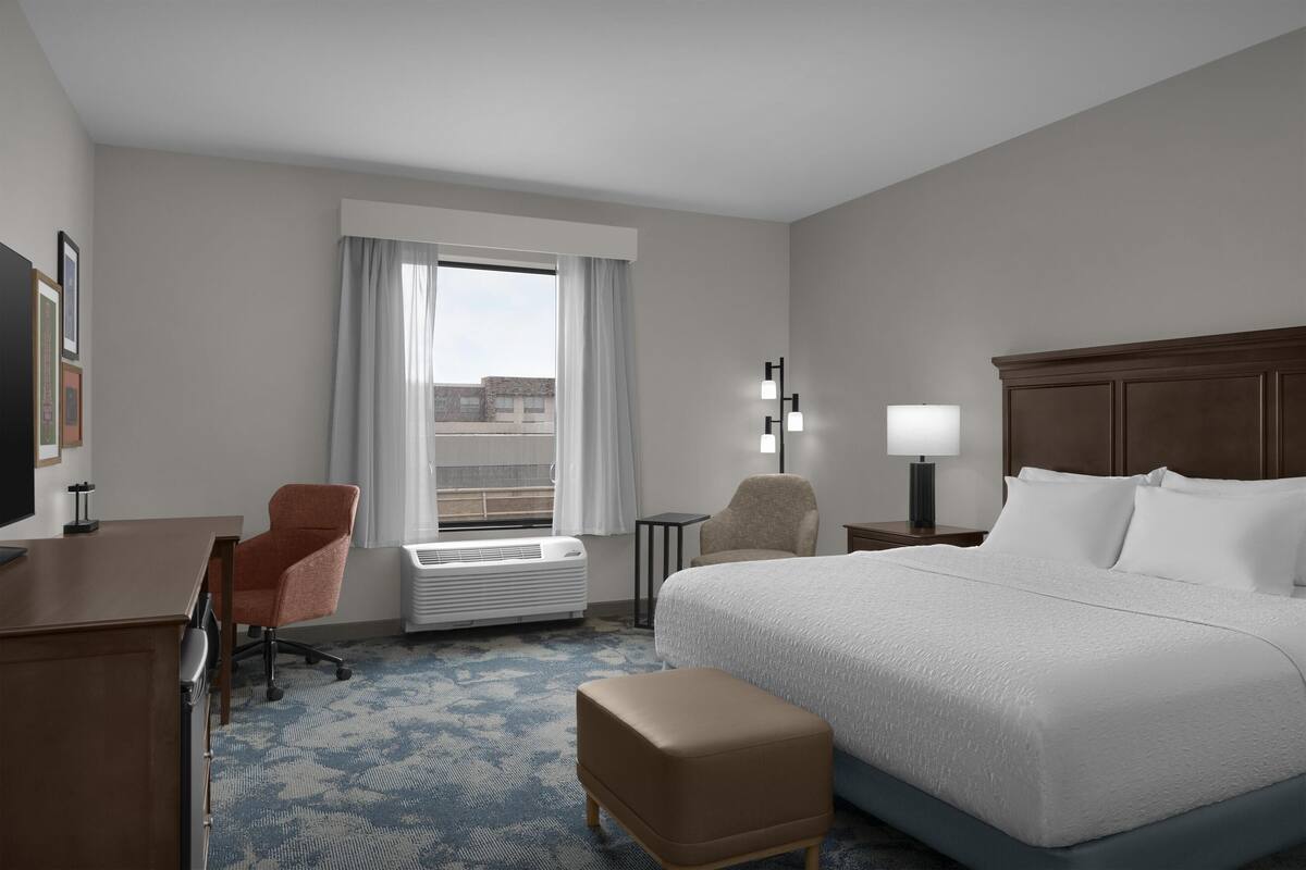 A hotel room with a bed, a desk, a chair, a lamp, and a window with curtains.
