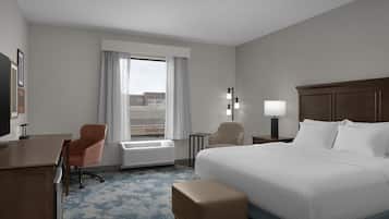 Habitació d'hotel amb llit, escriptori, cadira, làmpada i finestra amb cortines.