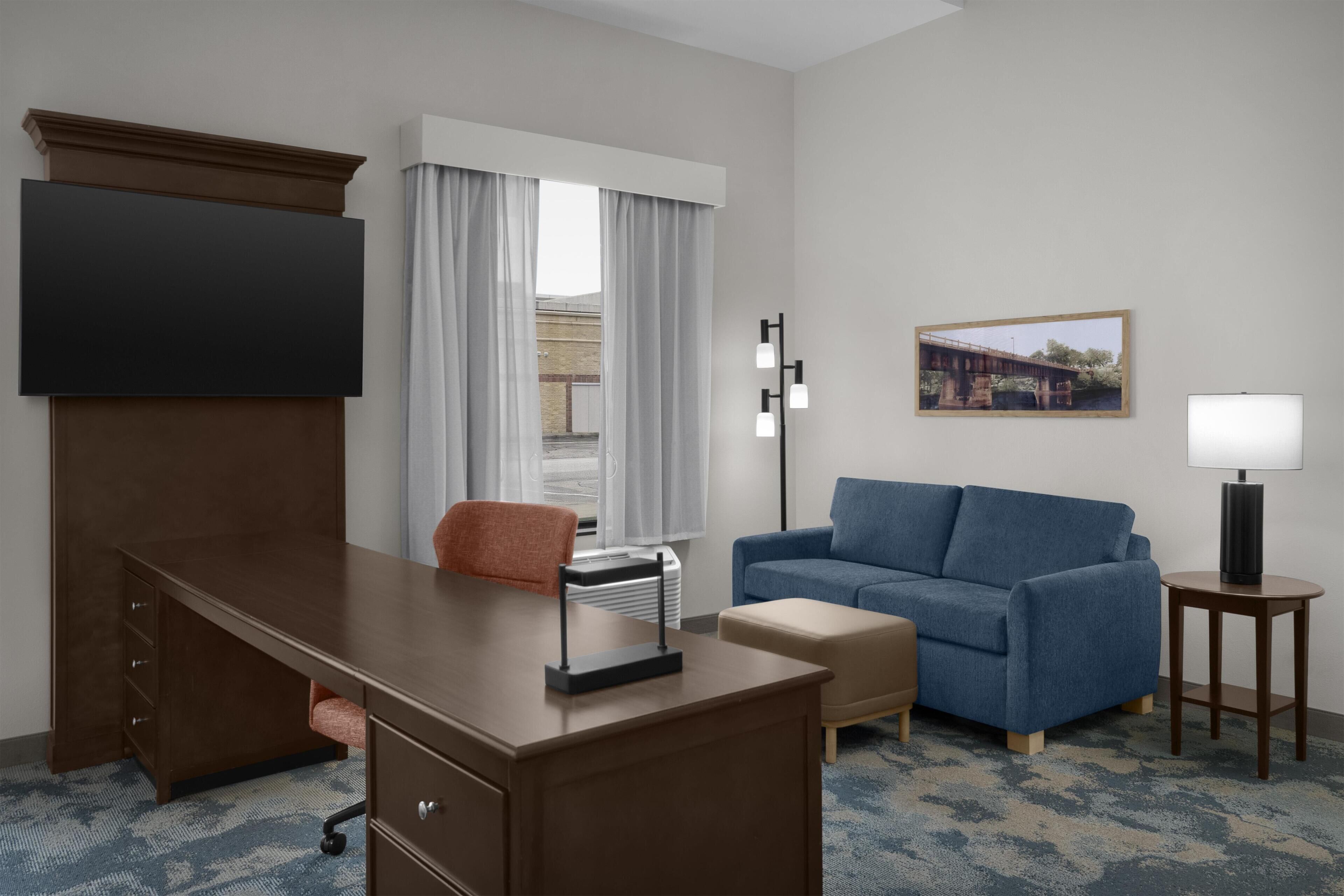 A hotel room with a desk, a sofa, a TV, a lamp, and a painting on the wall.