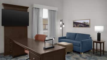 A hotel room with a desk, a sofa, a TV, a lamp, and a painting on the wall.