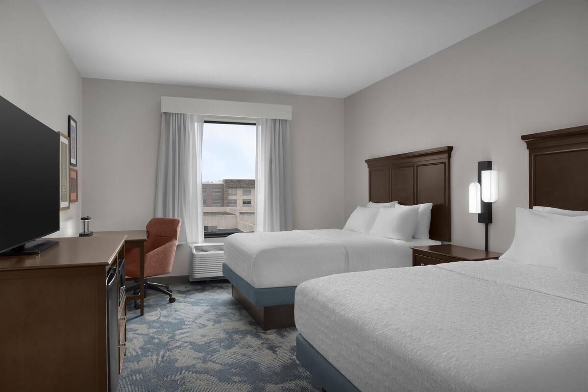 A hotel room with two beds, a desk, a chair, a television, and a window with curtains.