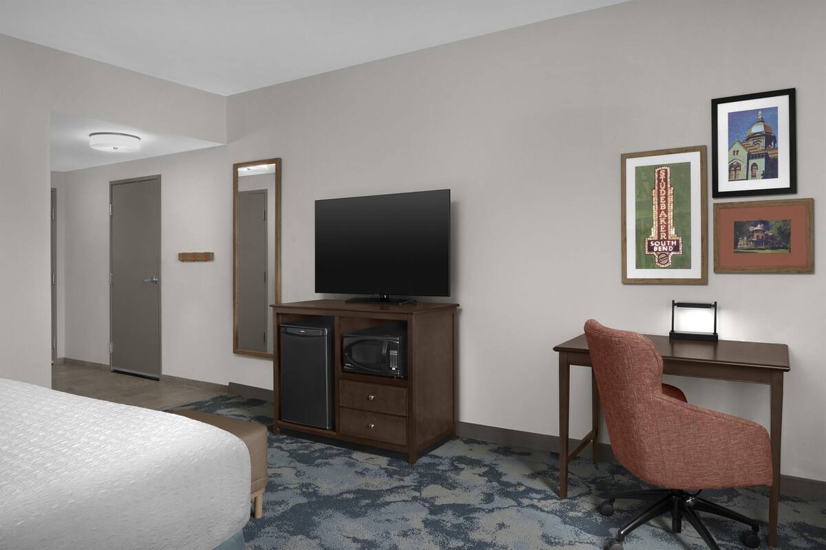 A hotel room with a bed, a TV, a desk with a chair, a microwave, a refrigerator, and framed pictures on the wall.