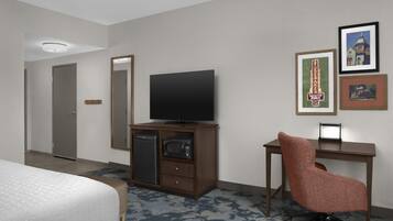 Una habitación de hotel con cama, televisión, escritorio con silla, microondas, refrigerador y cuadros en la pared.