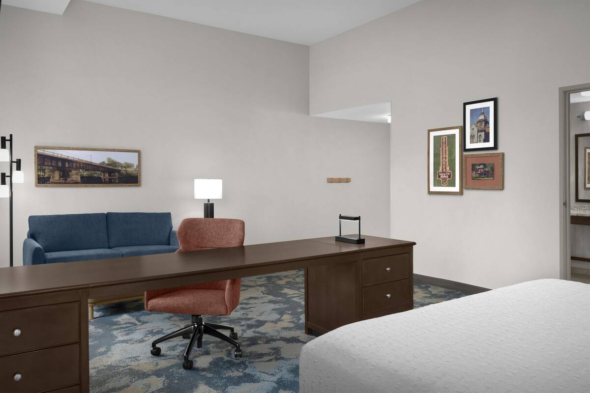A hotel room with a bed, a desk, a chair, a sofa, a lamp, and framed pictures on the wall.